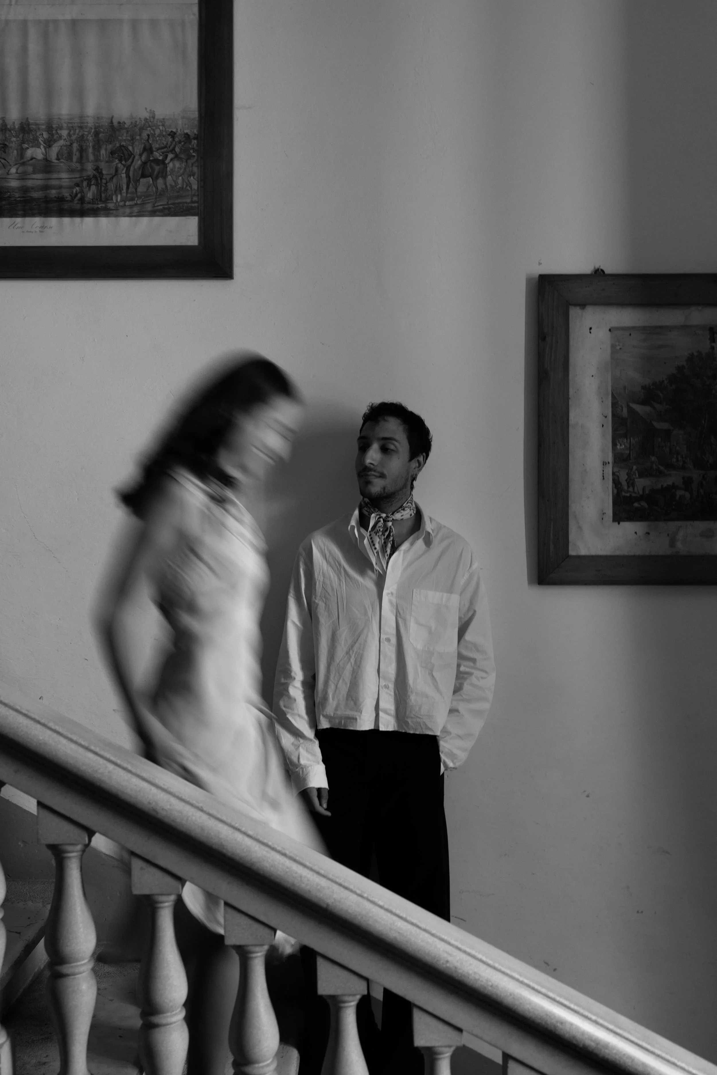 A young man with dark hair and a mustache standing near a wall, wearing a white shirt and a patterned scarf around his neck, while a woman with dark hair in motion, wearing a light-colored blouse, walks past him on a staircase in an indoor setting with framed artwork on the wall.