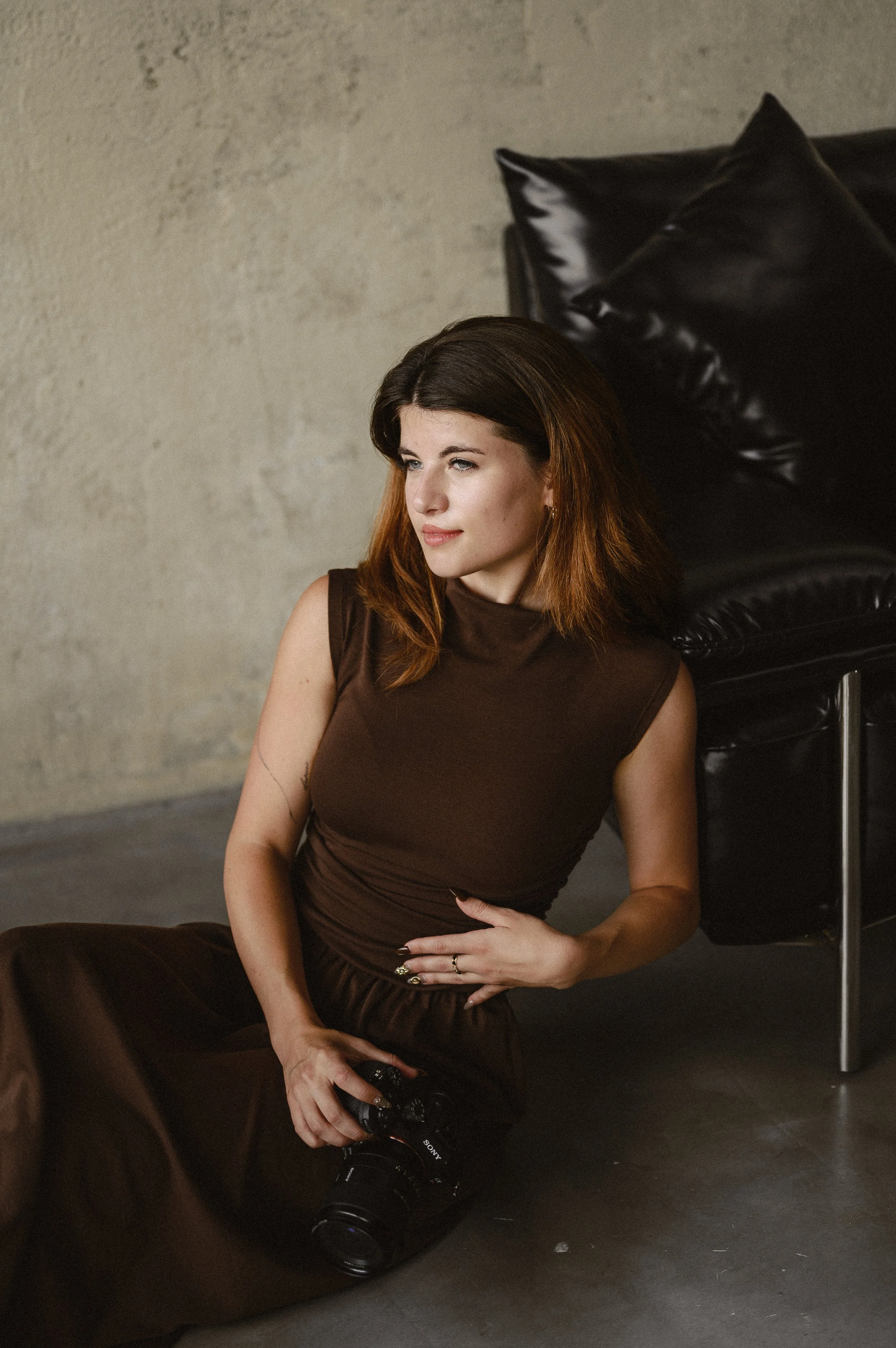 A woman with brown hair and blue eyes sitting on the floor, holding a camera, wearing a brown sleeveless dress, with black leather pillows and a beige textured wall in the background.