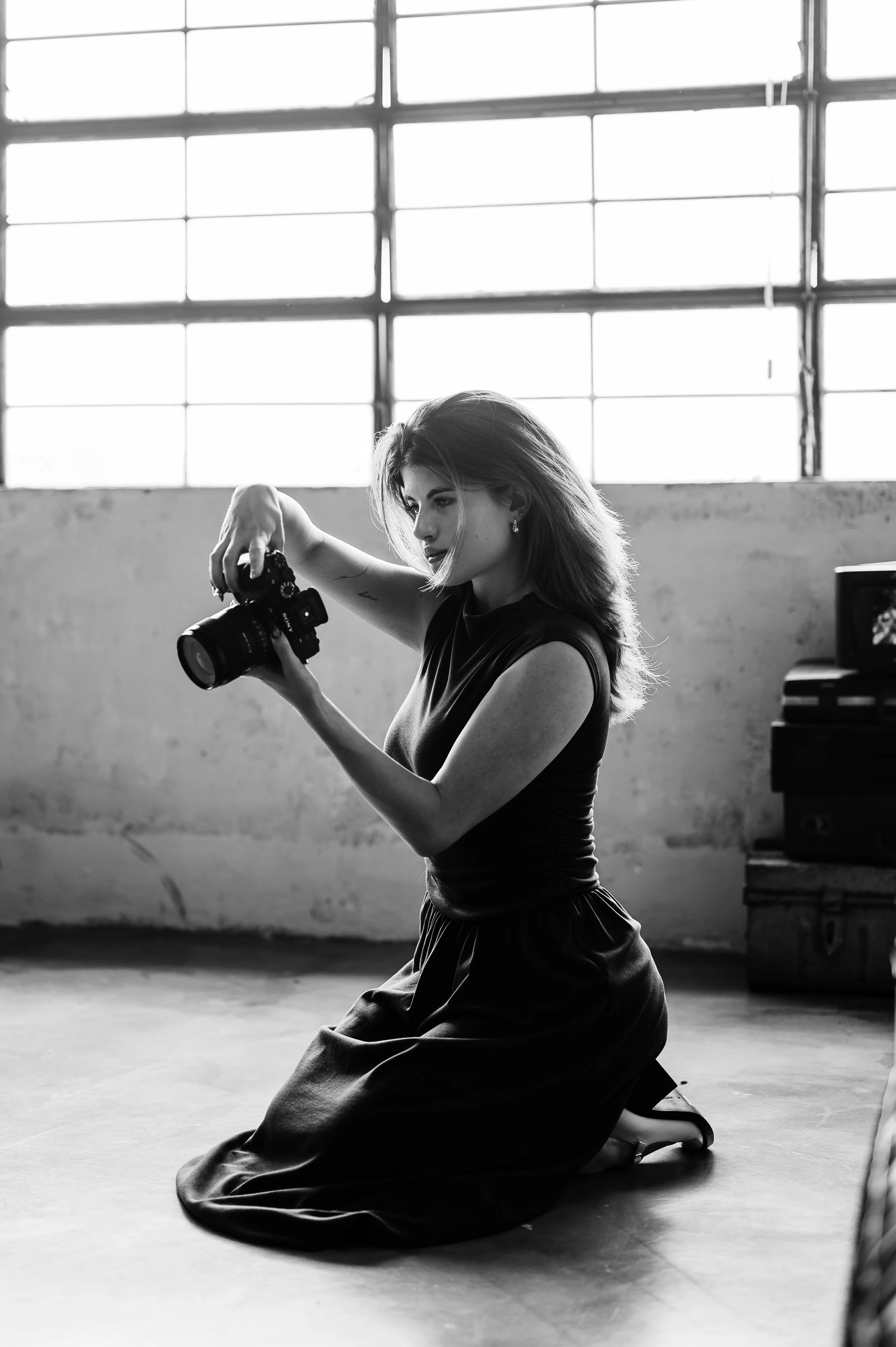 A young woman with shoulder-length hair and earrings is kneeling on the floor in a spacious, industrial-style room with large windows. She is holding a camera and appearing to take a photo, with natural light illuminating her profile.