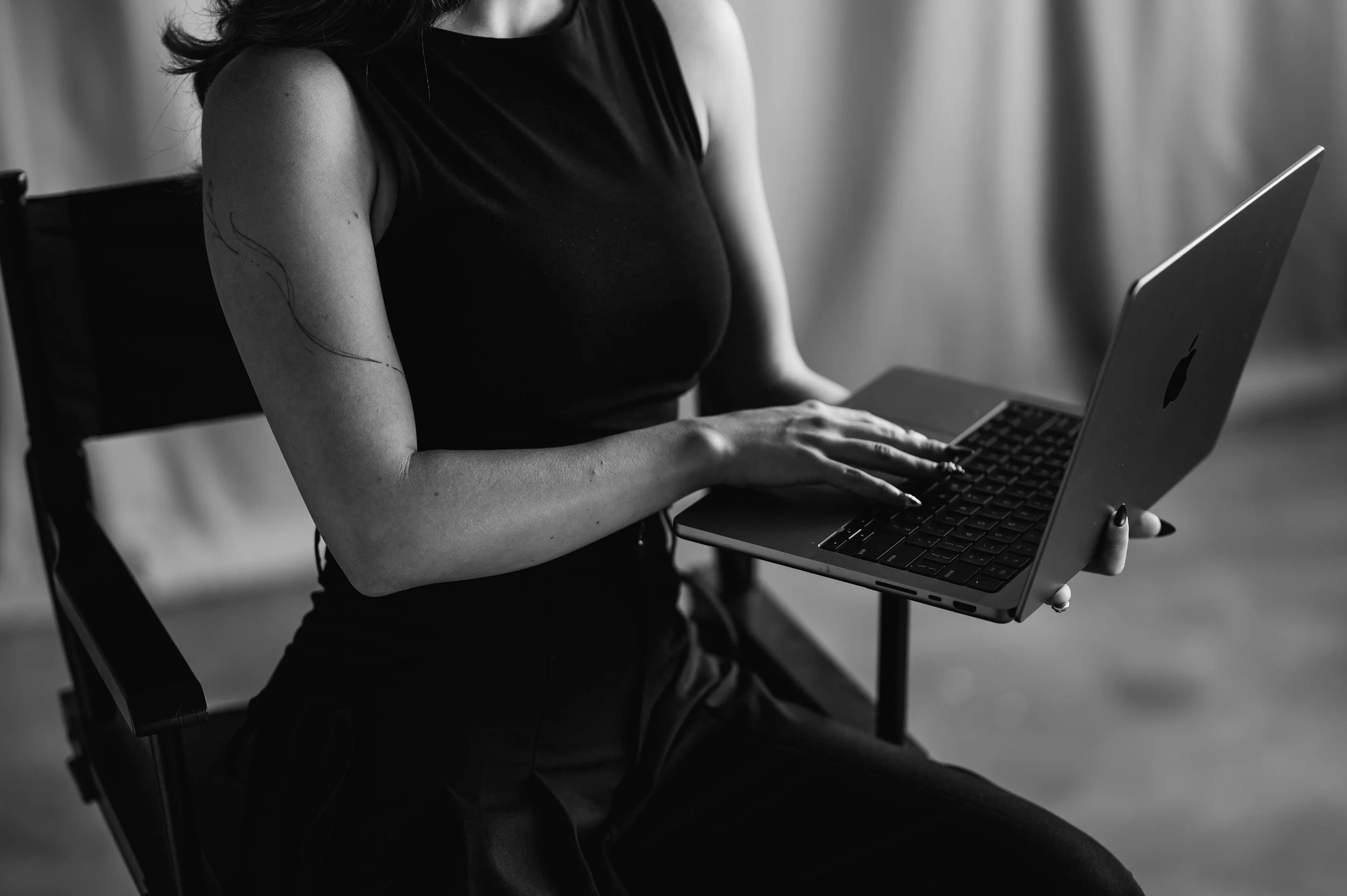 A woman sitting on a chair, holding an open MacBook laptop on her lap, in a black and white photograph.