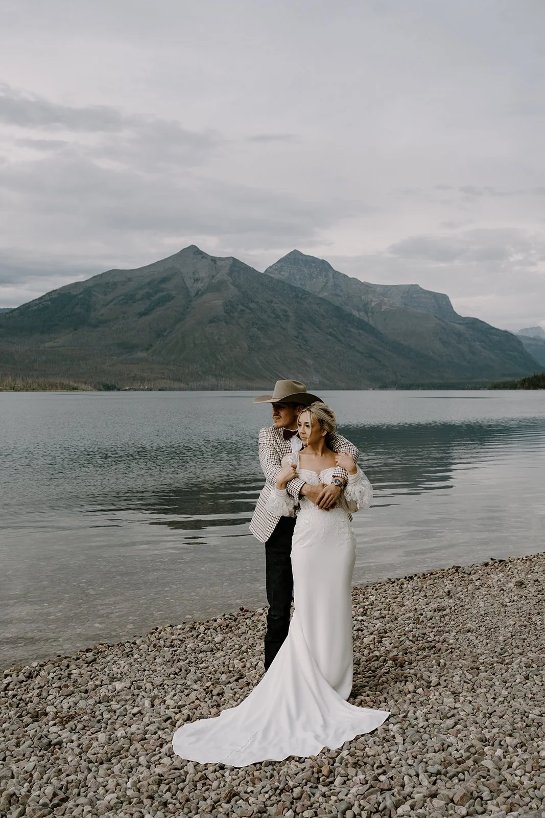 A bride and groom standing on a rocky lakeshore with mountains in the background. The groom is wearing a cowboy hat, checkered blazer, and black pants. The bride is in a white wedding dress with lace sleeves. They are embracing each other and looking into the distance.
