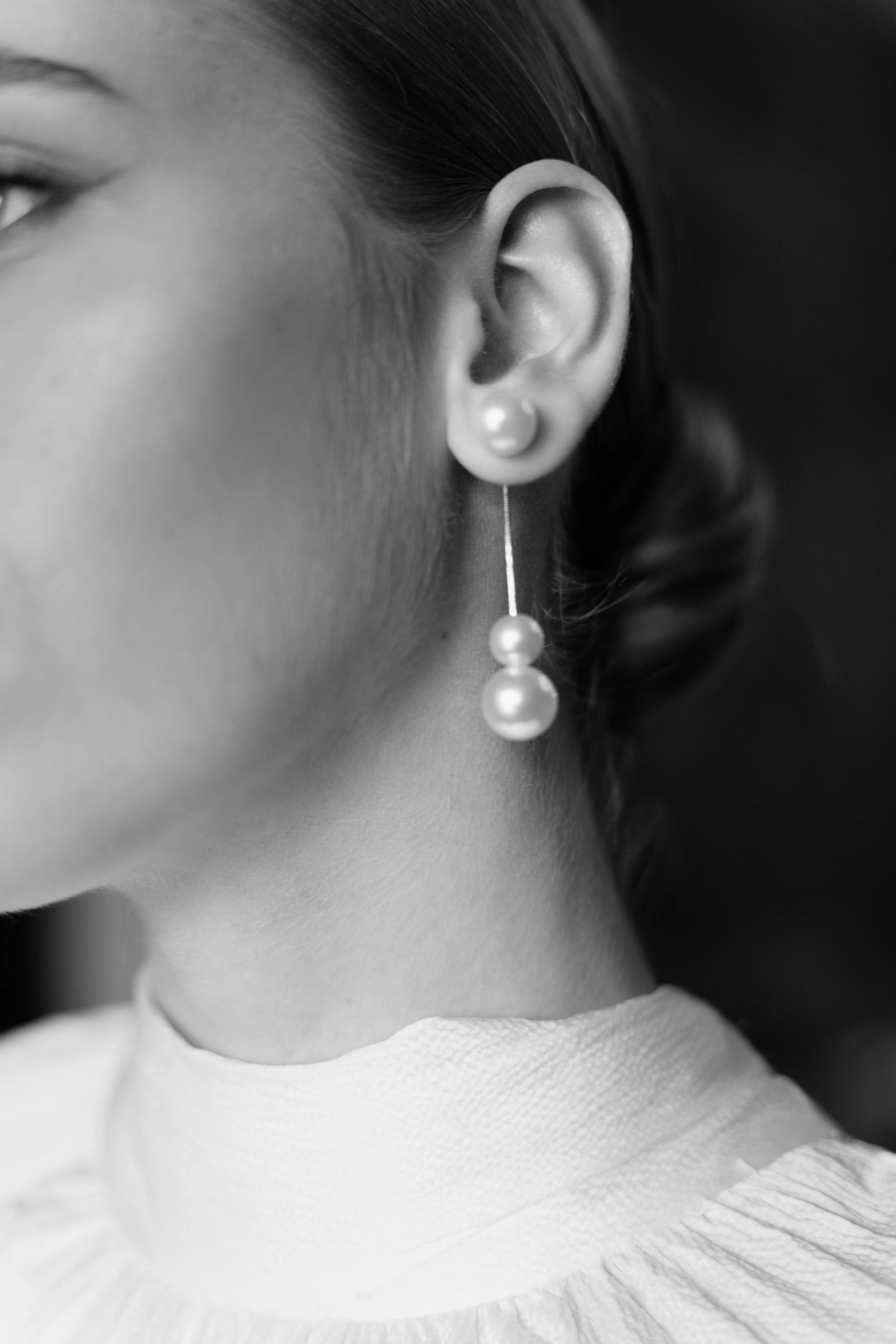 Close-up black and white photo of a woman's ear with a pearl earring and a portion of her face visible. The focus is on the ear and jewelry.
