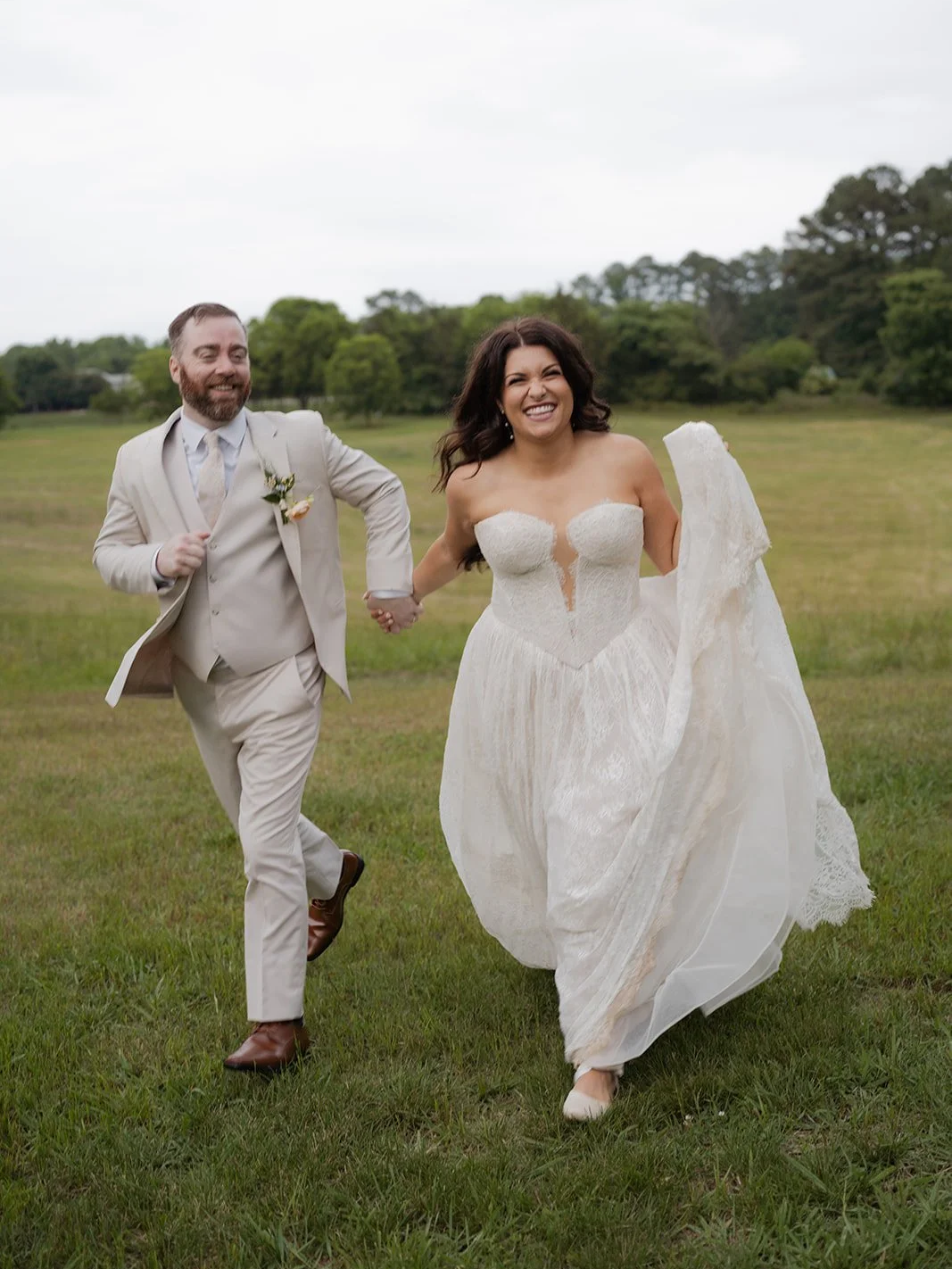 Bride and groom running hand in hand on a grassy field during their wedding, with trees and cloudy sky in the background.