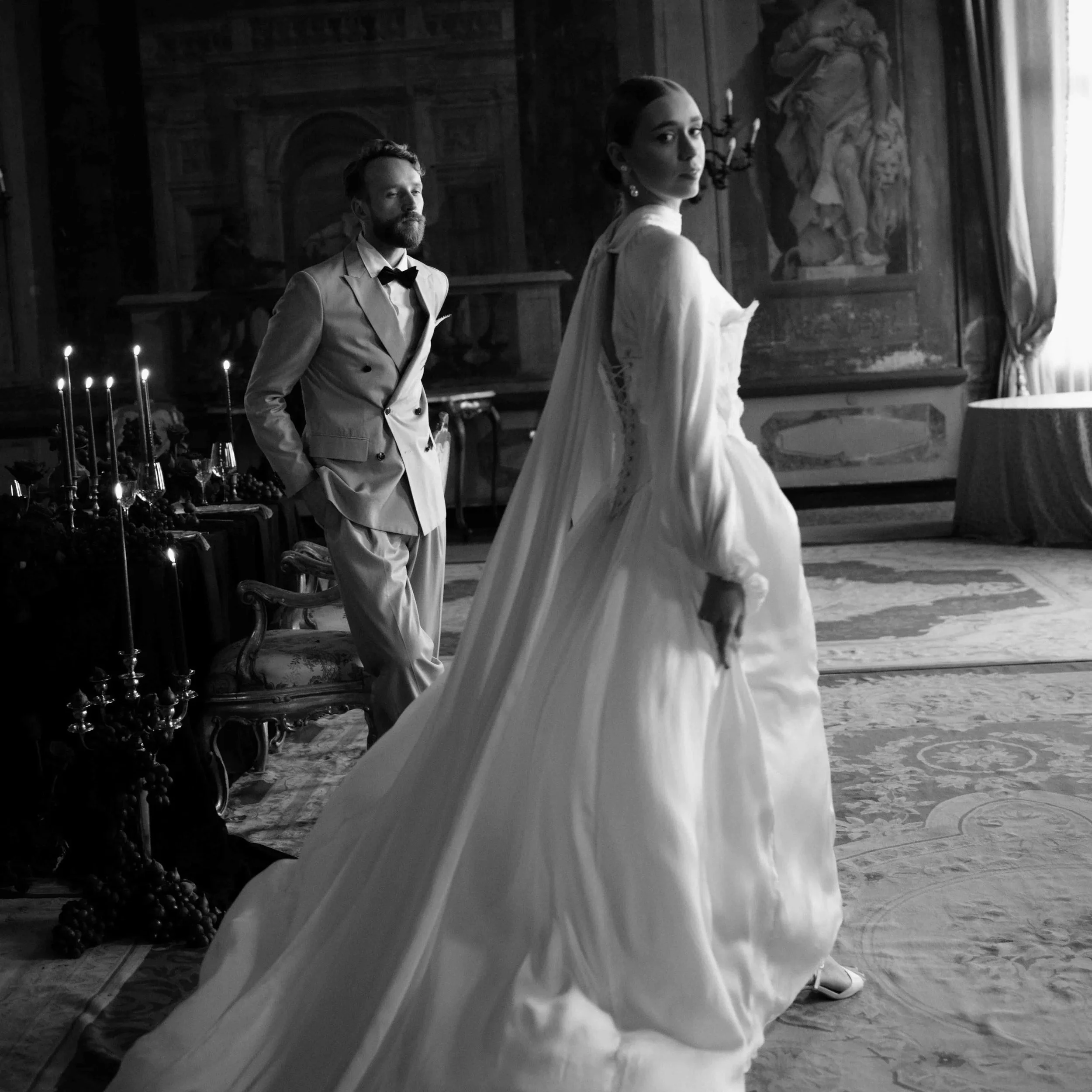 A black and white photograph of a man and a woman dressed in vintage formal attire standing in a grand room with ornate furniture and paintings. The man has a beard and is wearing a tuxedo, while the woman has her hair styled up and is wearing a white dress.