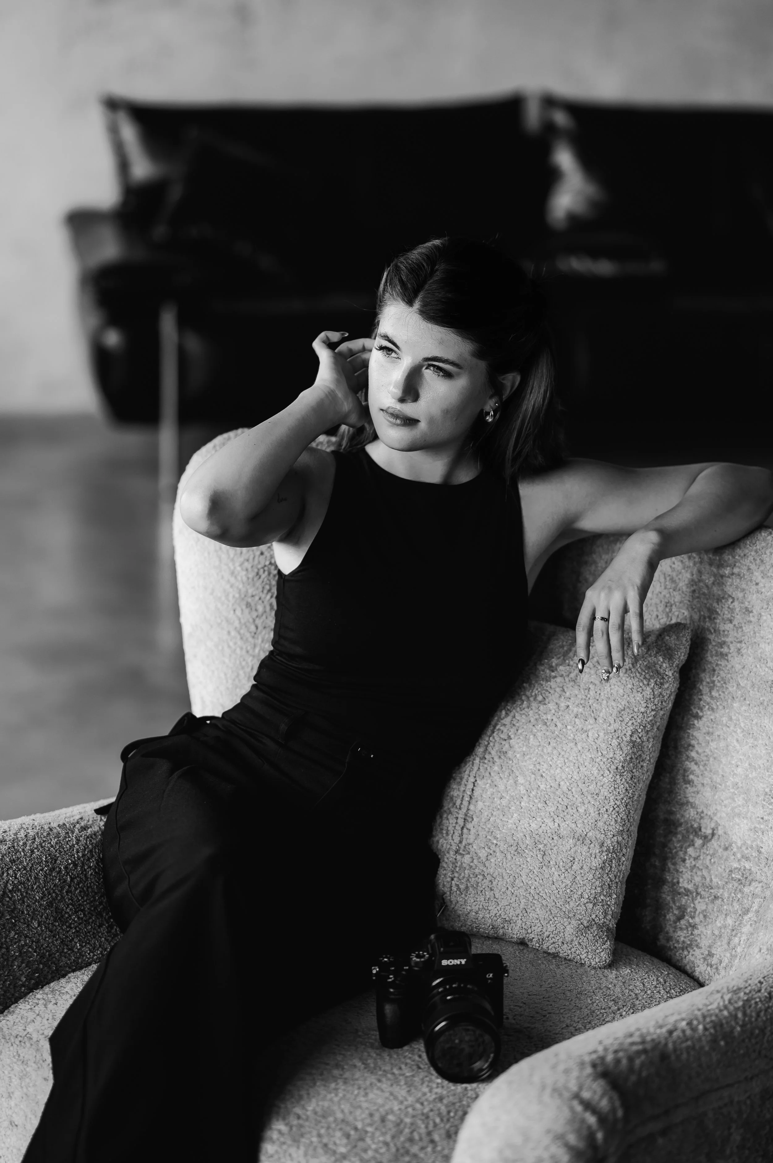 Black and white photo of a woman with dark hair, sitting on a textured armchair, with a Sony camera resting on the seat next to her.