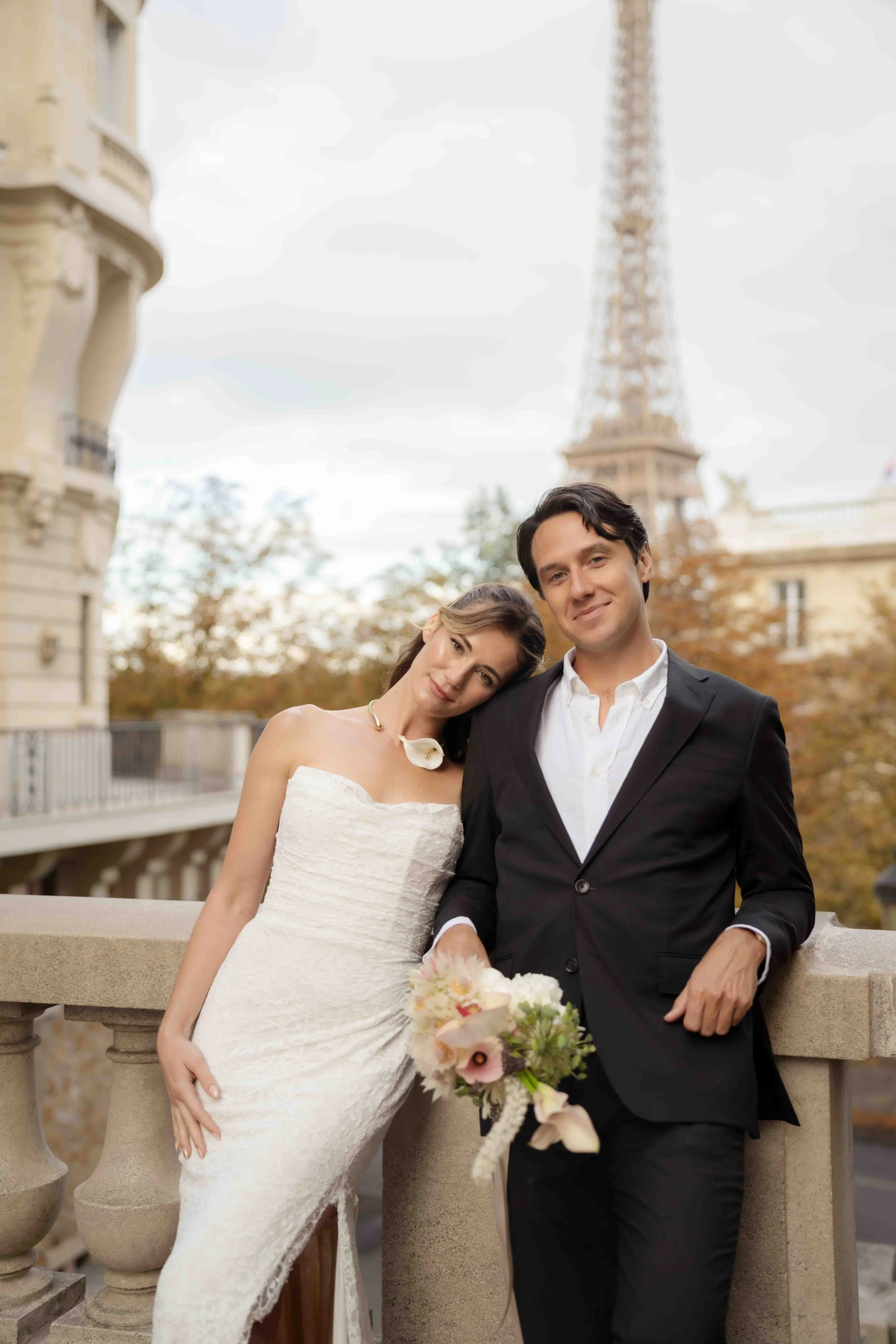 A bride and groom standing close together on a Paris balcony with the Eiffel Tower in the background. The bride leans her head on the groom's shoulder, holding a bouquet of flowers. The groom is smiling and wearing a black suit and white shirt.