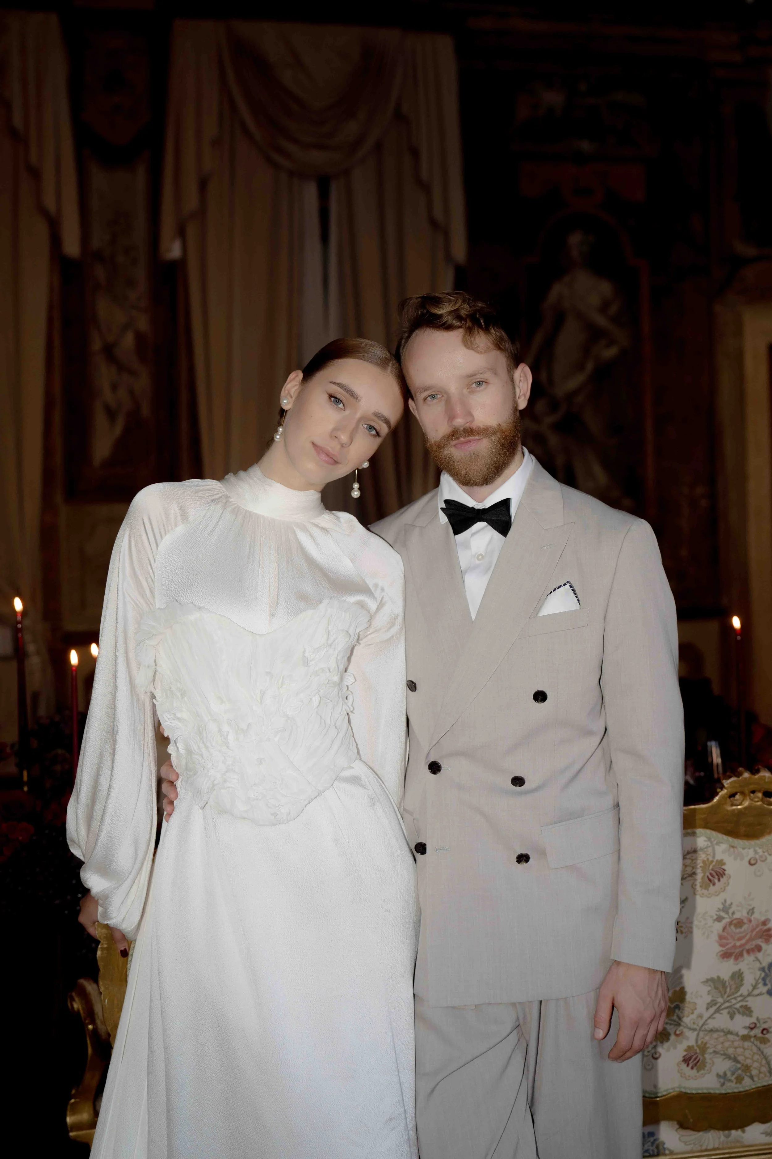 A man and woman in formal wedding attire standing close together in a richly decorated room. The woman is wearing a white wedding dress with pearl earrings, and the man is dressed in a light-colored tuxedo with a black bow tie.