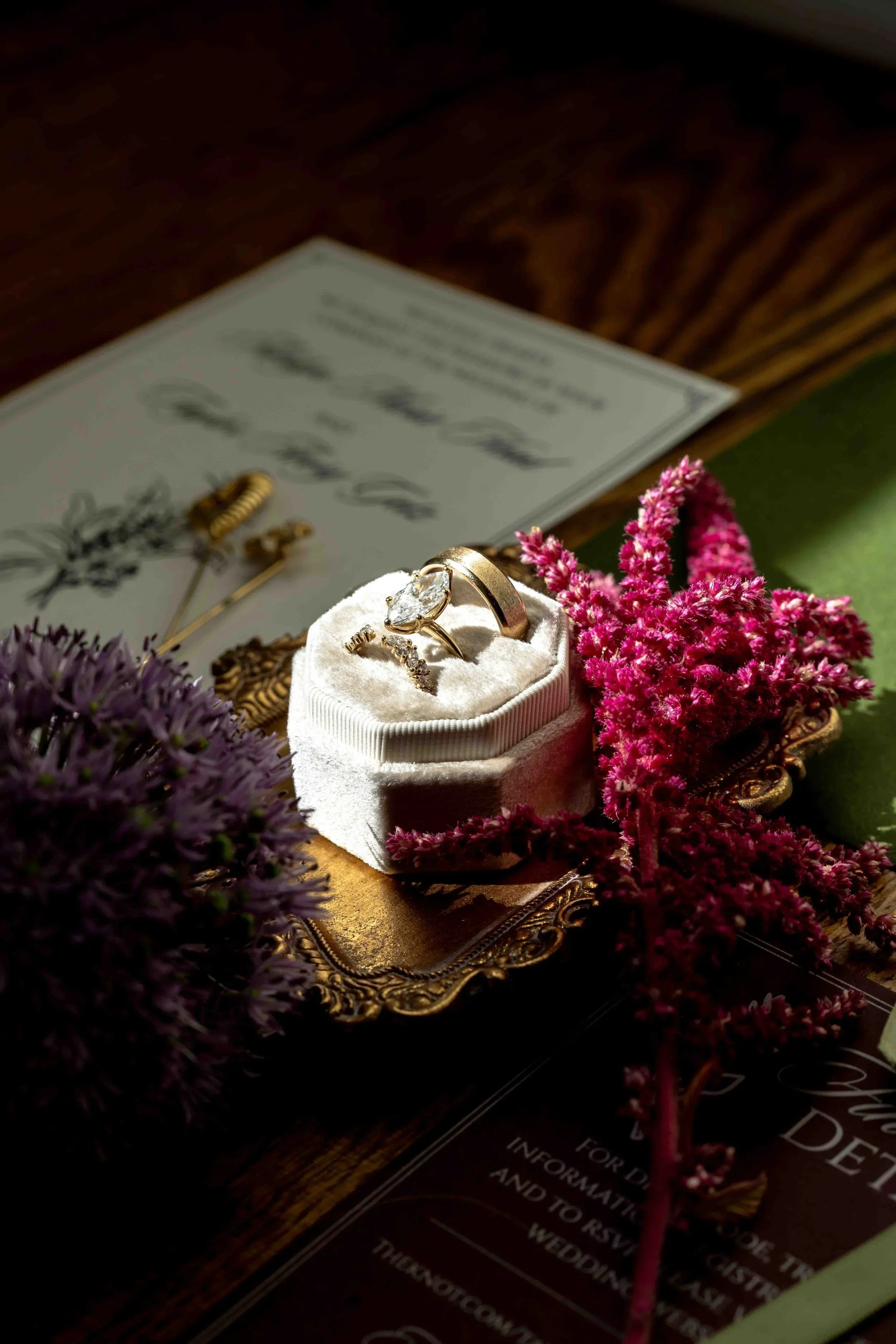 Close-up of a wedding ring set with a large center diamond on a white ring box. Surrounding flowers are pink and purple, and a blurred certificate or document is in the background on a dark wooden surface.