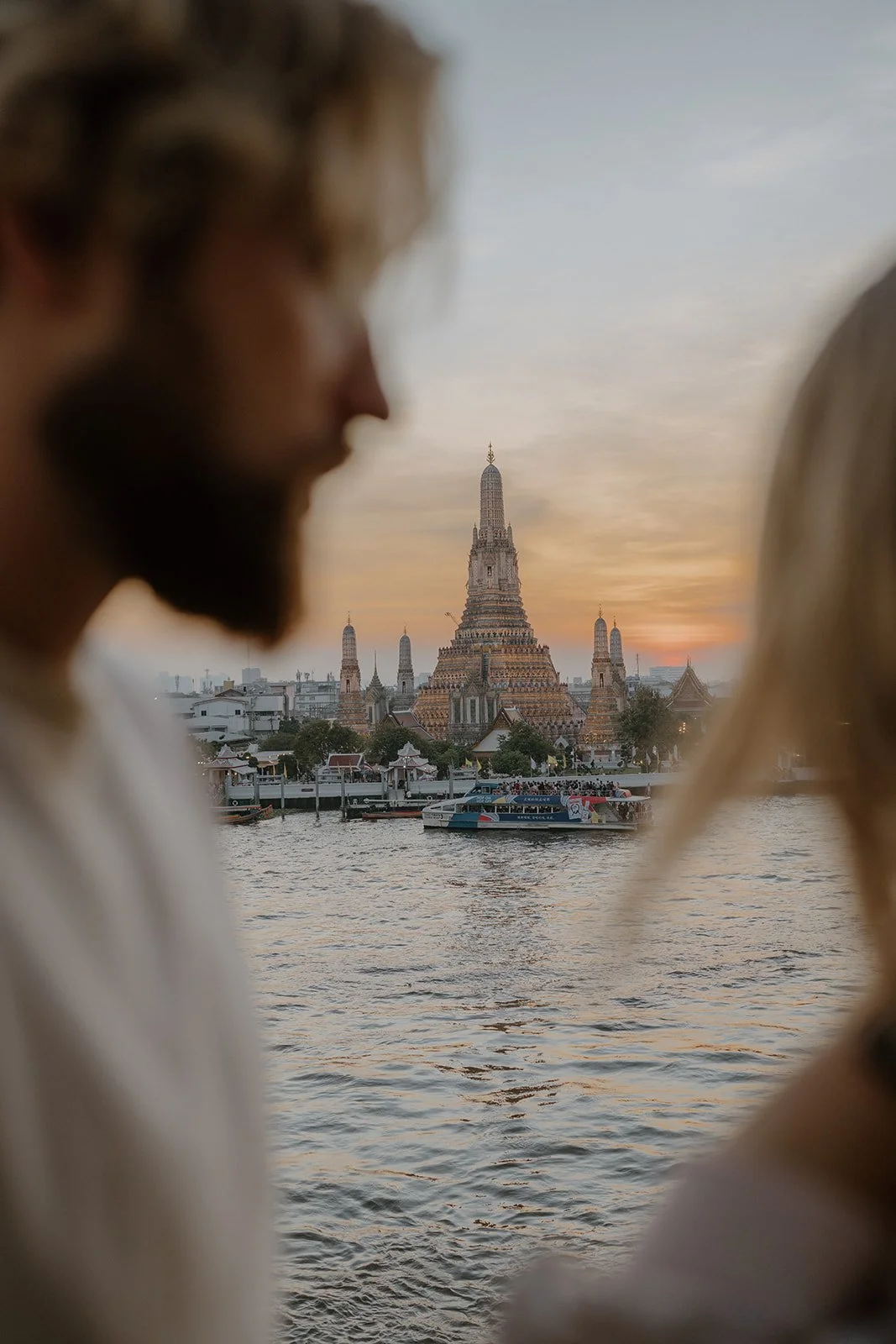 Blurred profile of a man with a beard looking towards a river, with a historic temple and sunset in the background.