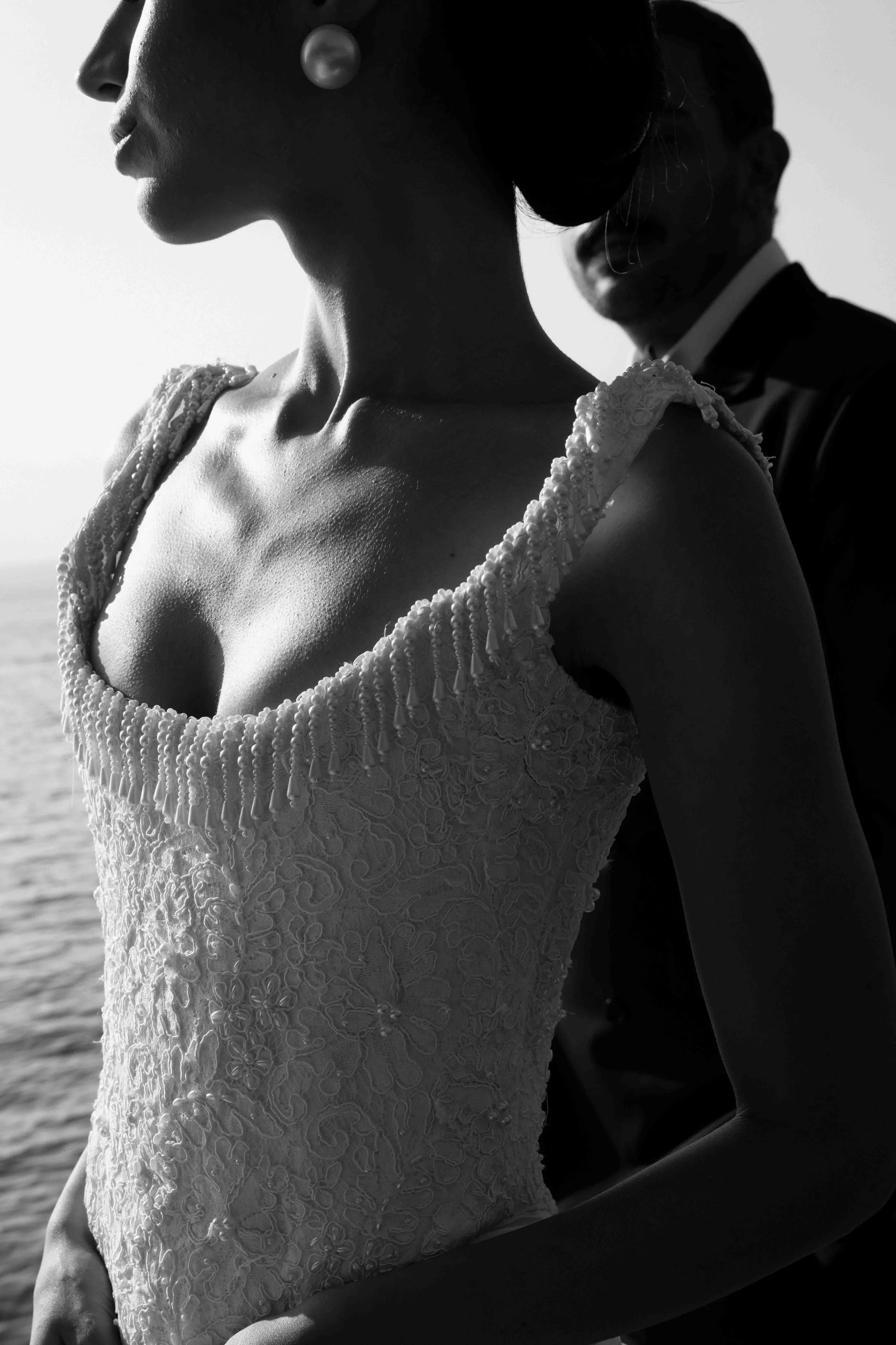 A black-and-white photo of a woman in a lace dress with beaded details, wearing a pearl earring, with two men in formal attire, one of whom is smiling, in the background.
