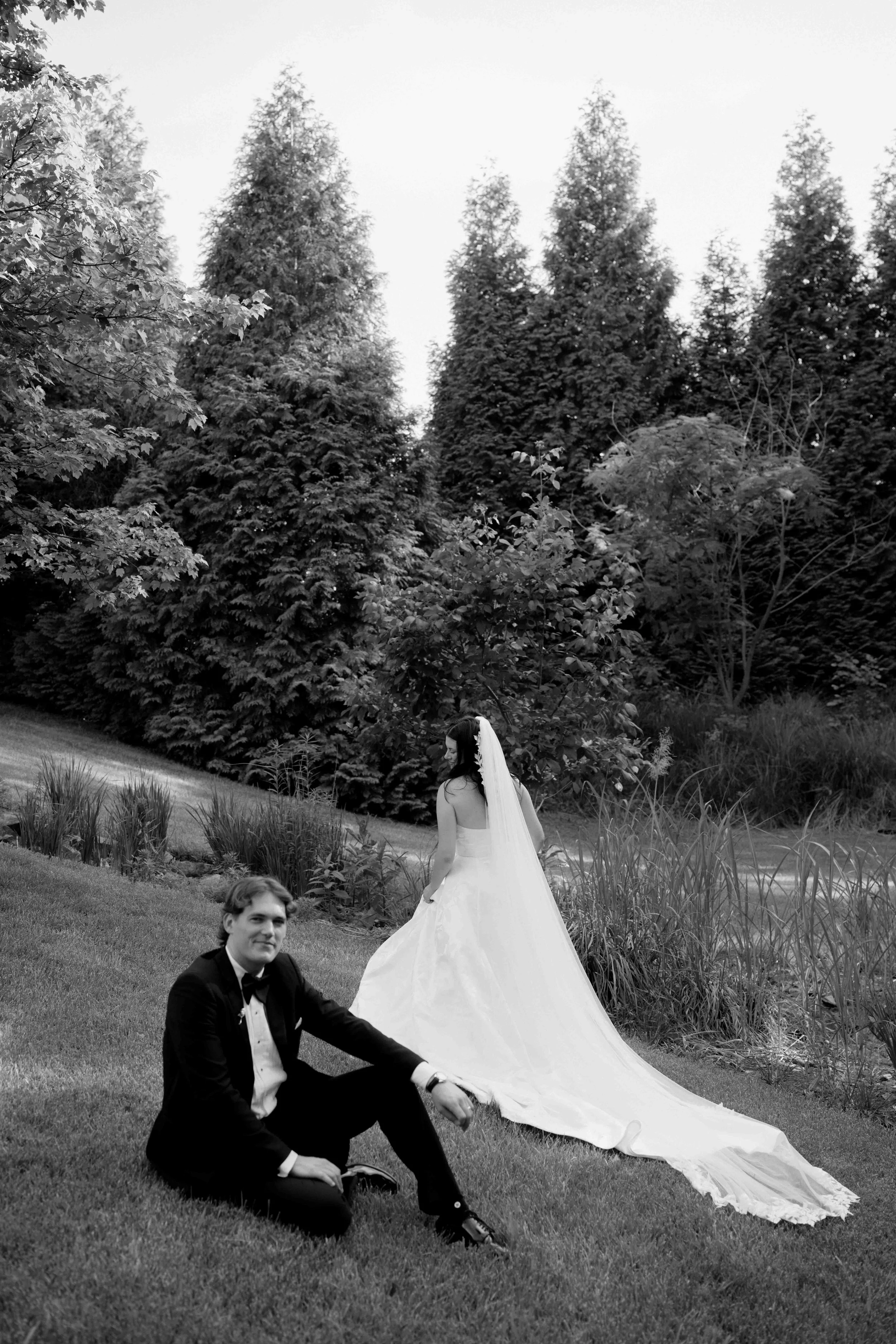 A black and white photo of a bride in a wedding dress with long train standing next to a groom in a tuxedo with bow tie, sitting on the grass in a garden with tall trees in the background.