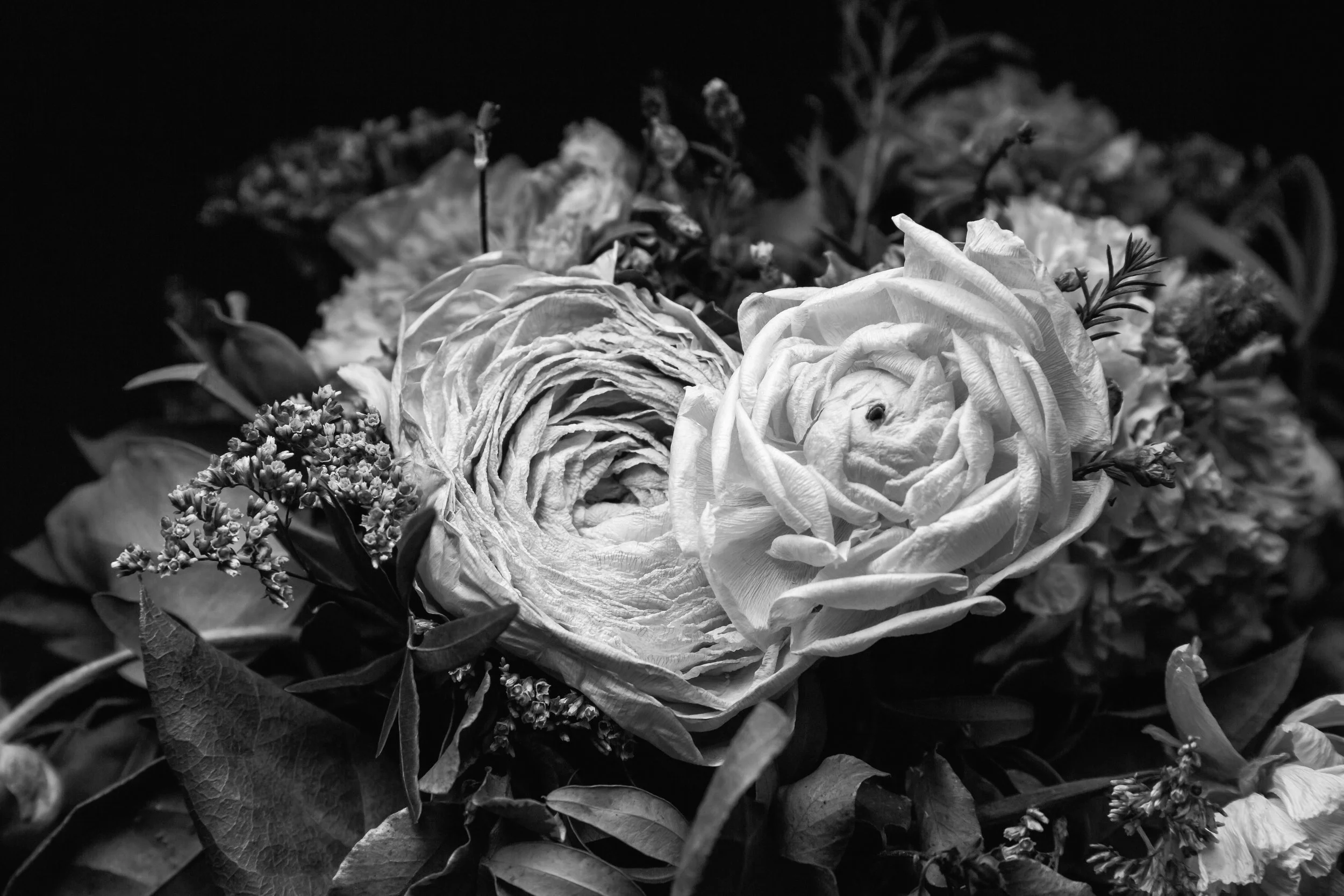A black and white close-up of a bouquet of flowers, including roses, with some wilted or dried petals and small filler flowers.