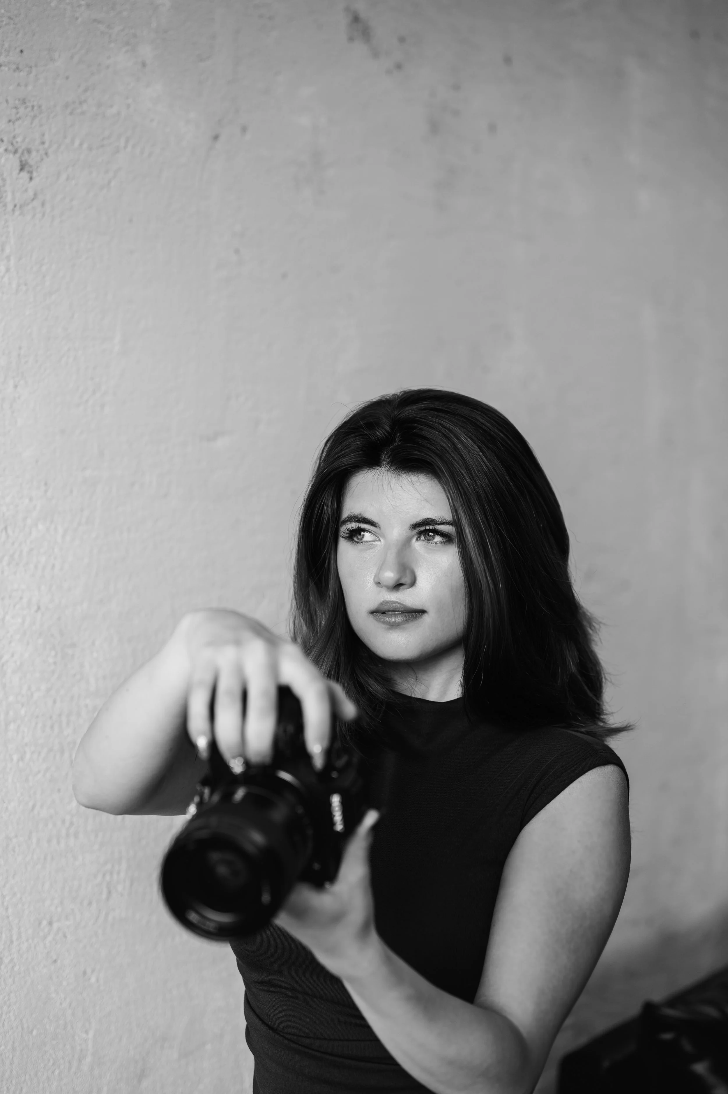 Black-and-white photo of a woman with dark hair taking a selfie with a camera, pointing it toward a mirror or reflective surface. She is wearing a sleeveless top and looking slightly to her left with a neutral expression, against a textured wall background.