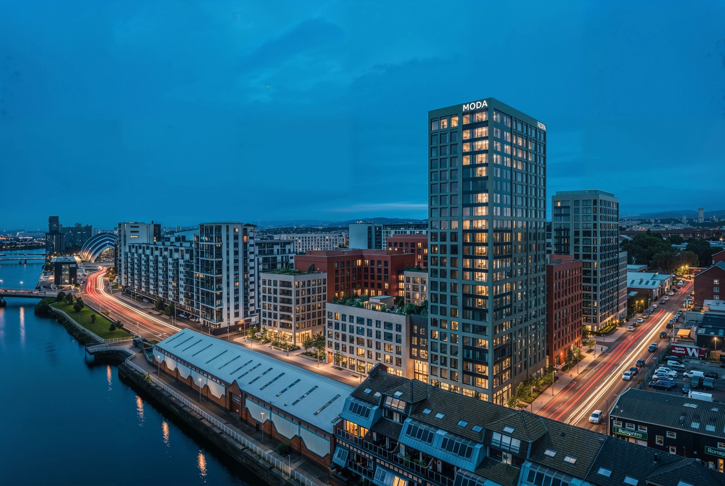 A cityscape at dusk featuring a tall modern building with illuminated windows and the word "MODA" on top. Surrounding buildings and a river with a pathway running alongside are visible, with light trails from moving vehicles on the streets.