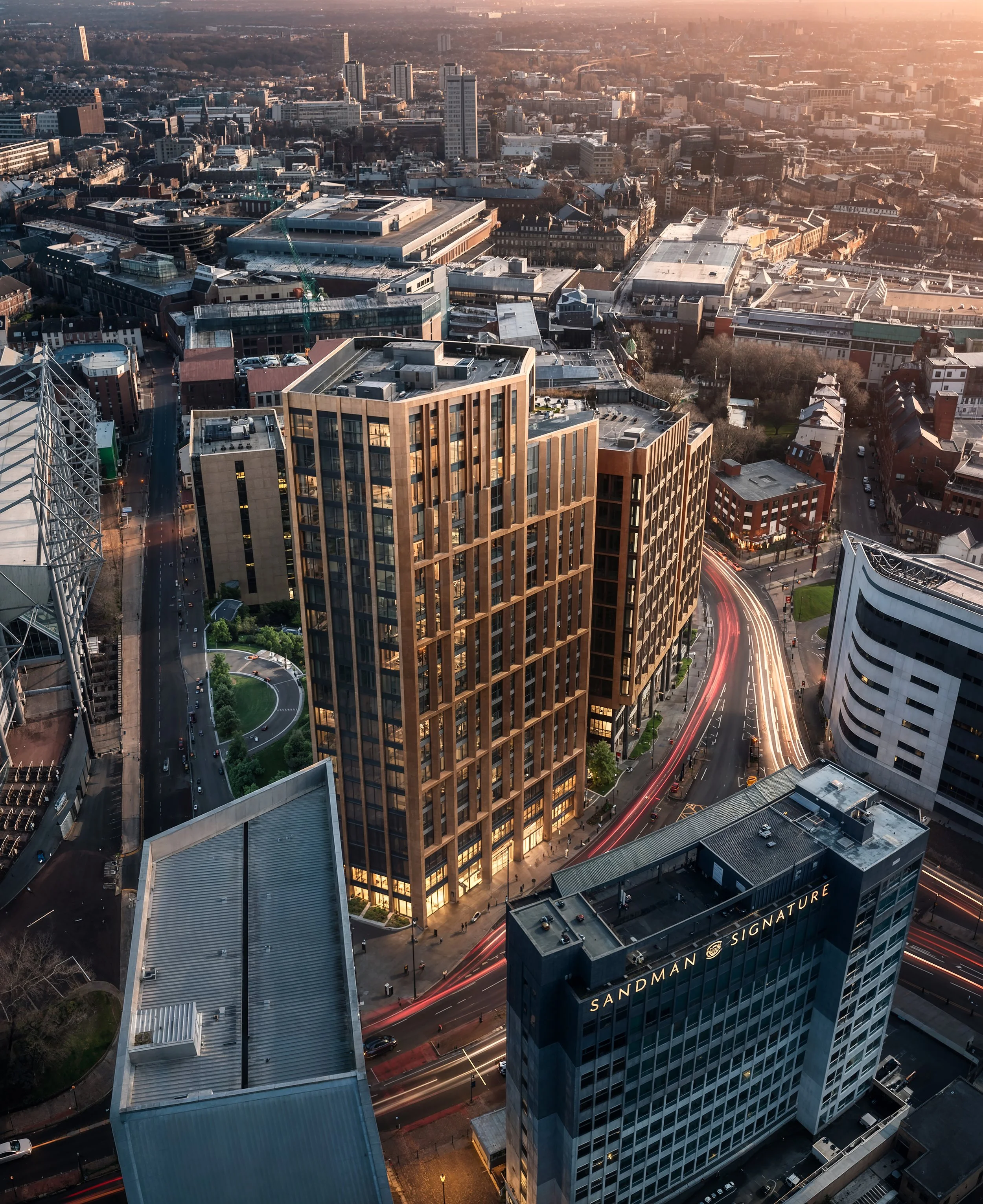 Aerial cityscape of a downtown area during sunset, featuring tall modern buildings, busy streets with light trails from moving vehicles, and a mix of architectural styles including glass and brick structures.