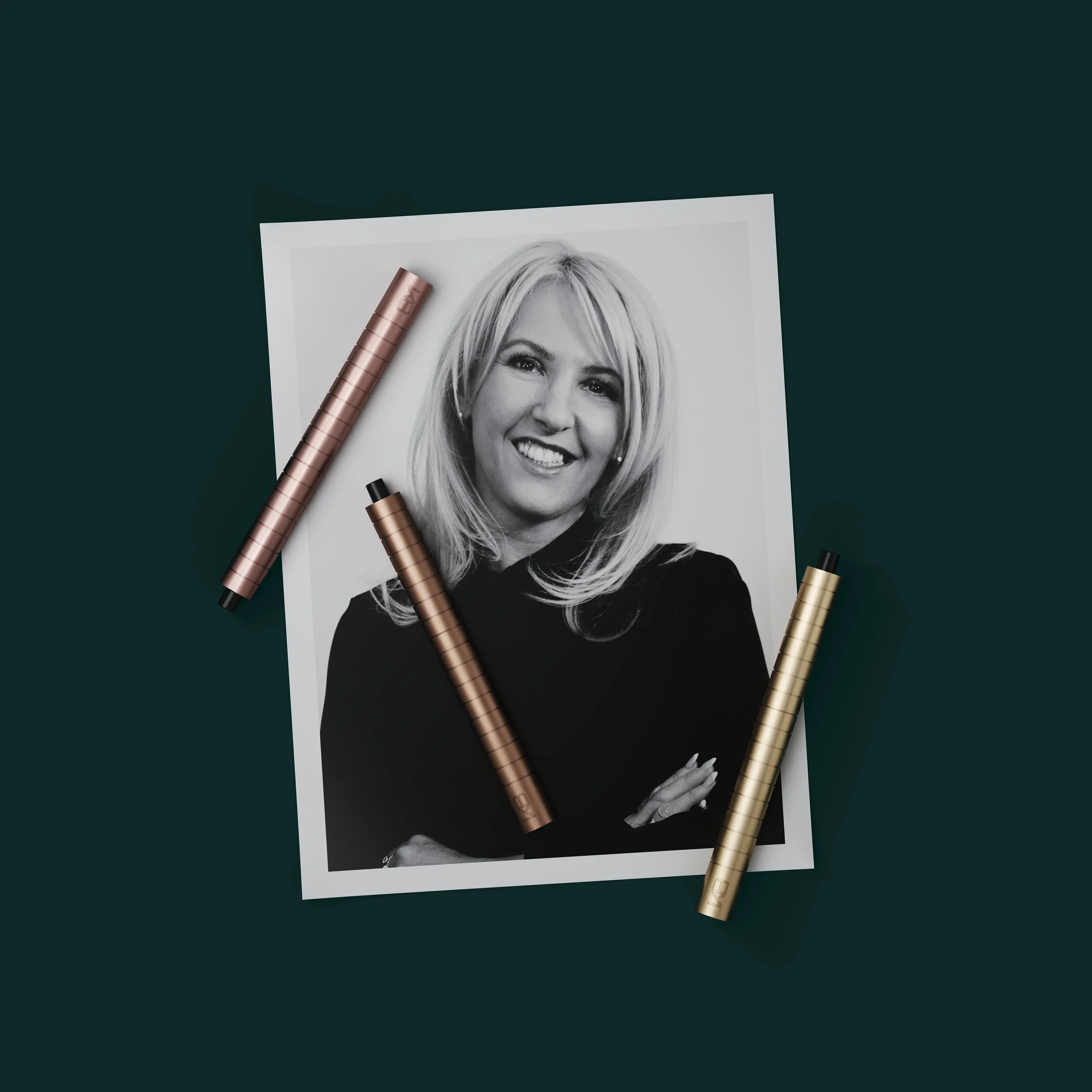 Black and white portrait photograph of a smiling woman with shoulder-length hair, surrounded by art markers on a dark green background.
