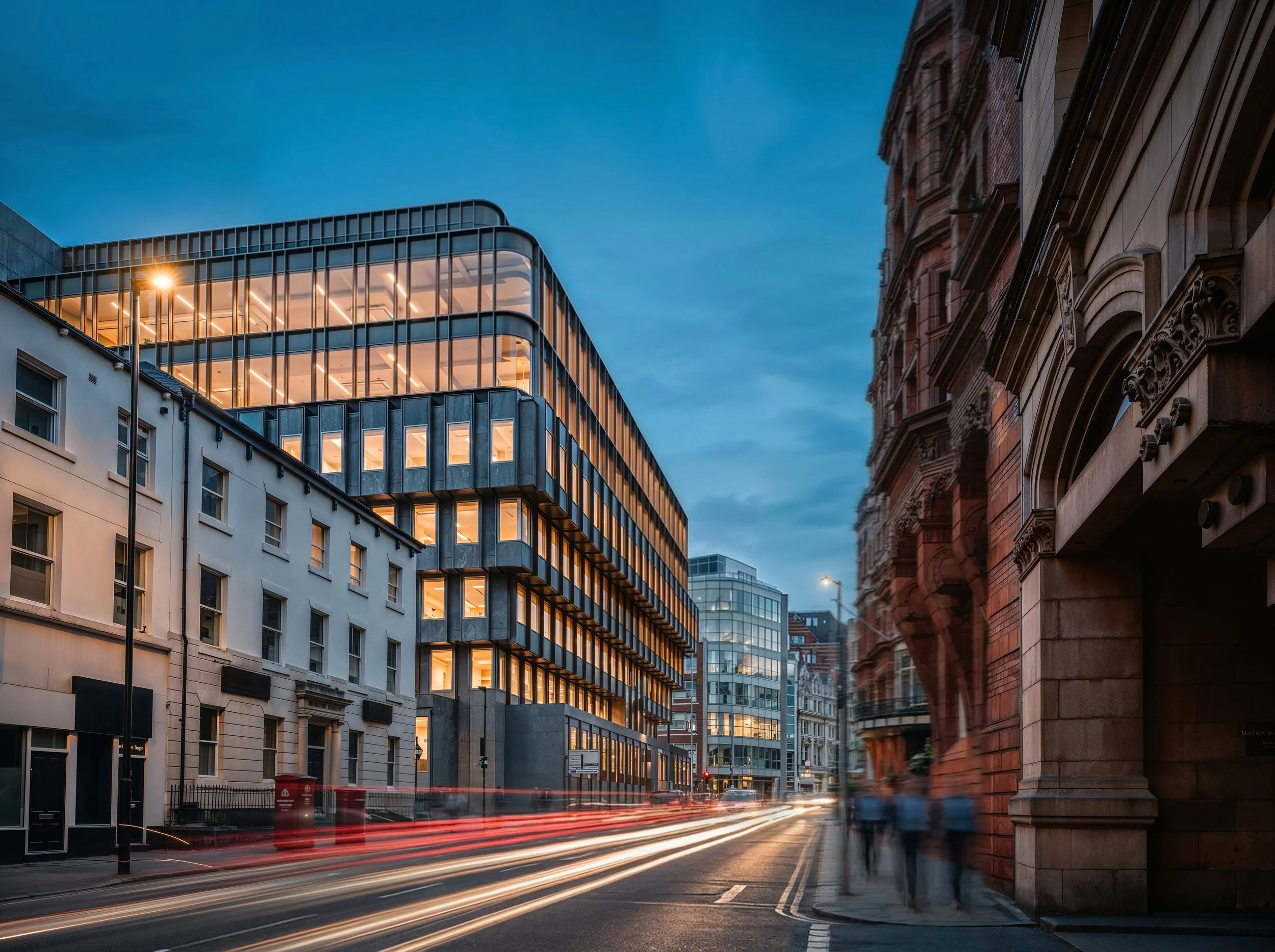 City street at dusk with modern glass building, historic brick buildings, and light trails from moving cars.