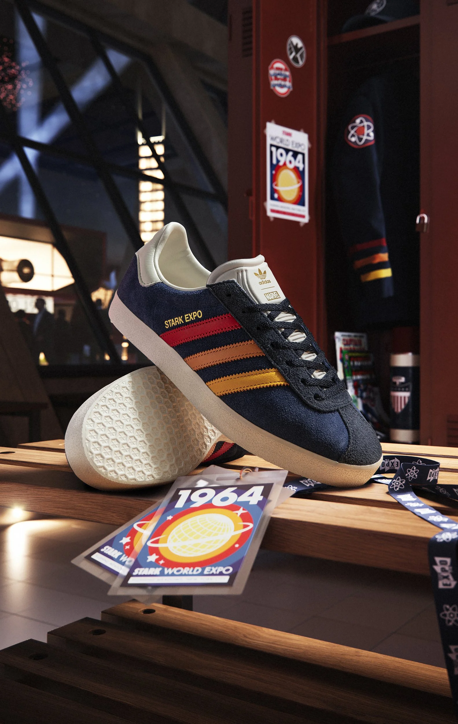 A sneaker displayed on a wooden surface with a poster in the background that says "1964 Stark World Expo" featuring a yellow planet with rings and stars. The sneaker is navy blue with red, orange, and yellow stripes and the words "Stark Expo" on the 