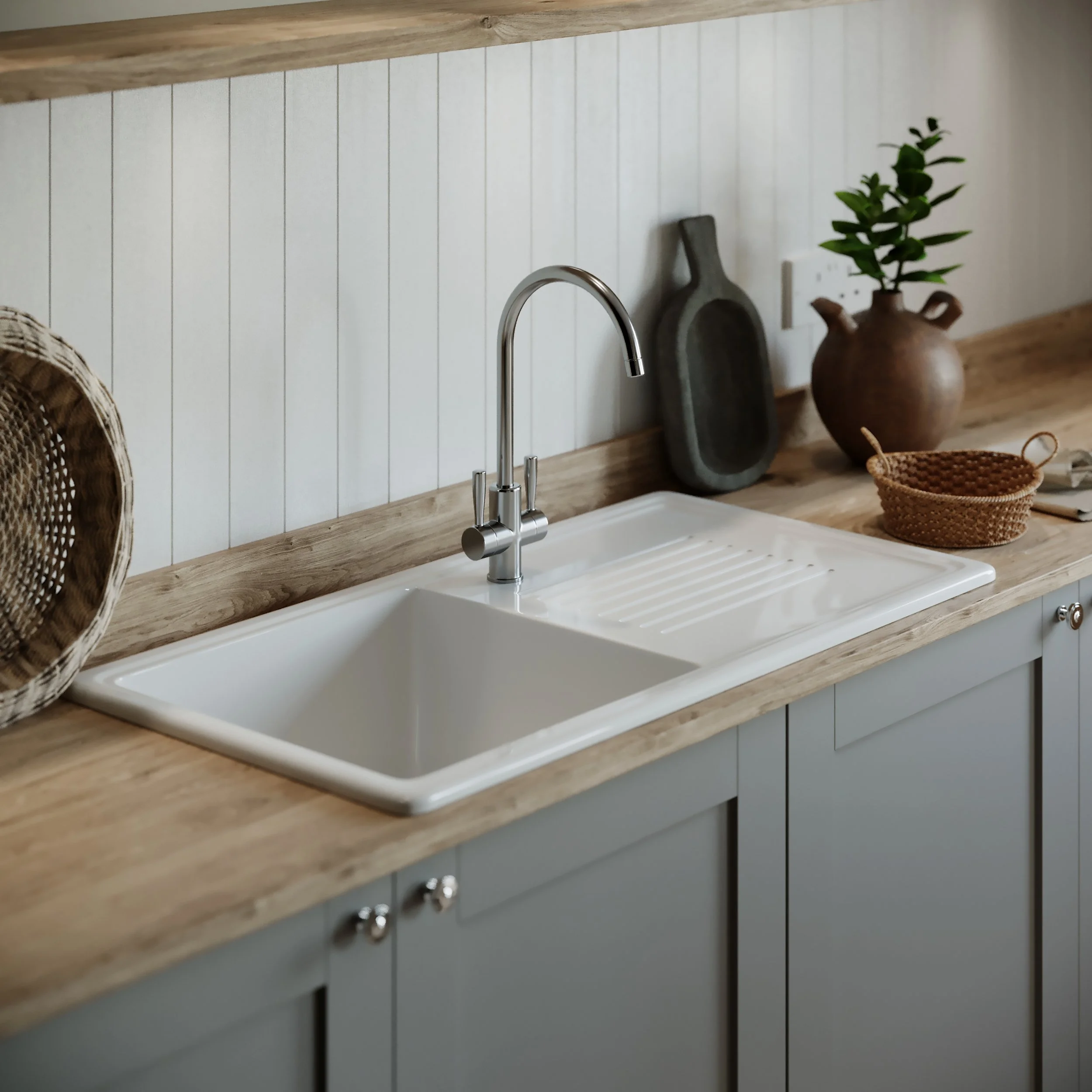 Kitchen countertop with a white sink, a silver faucet, a wicker basket on the left, a dark gray cutting board and a plant in a brown vase on the right, with a light-colored wooden shelf above and a white beadboard backsplash.