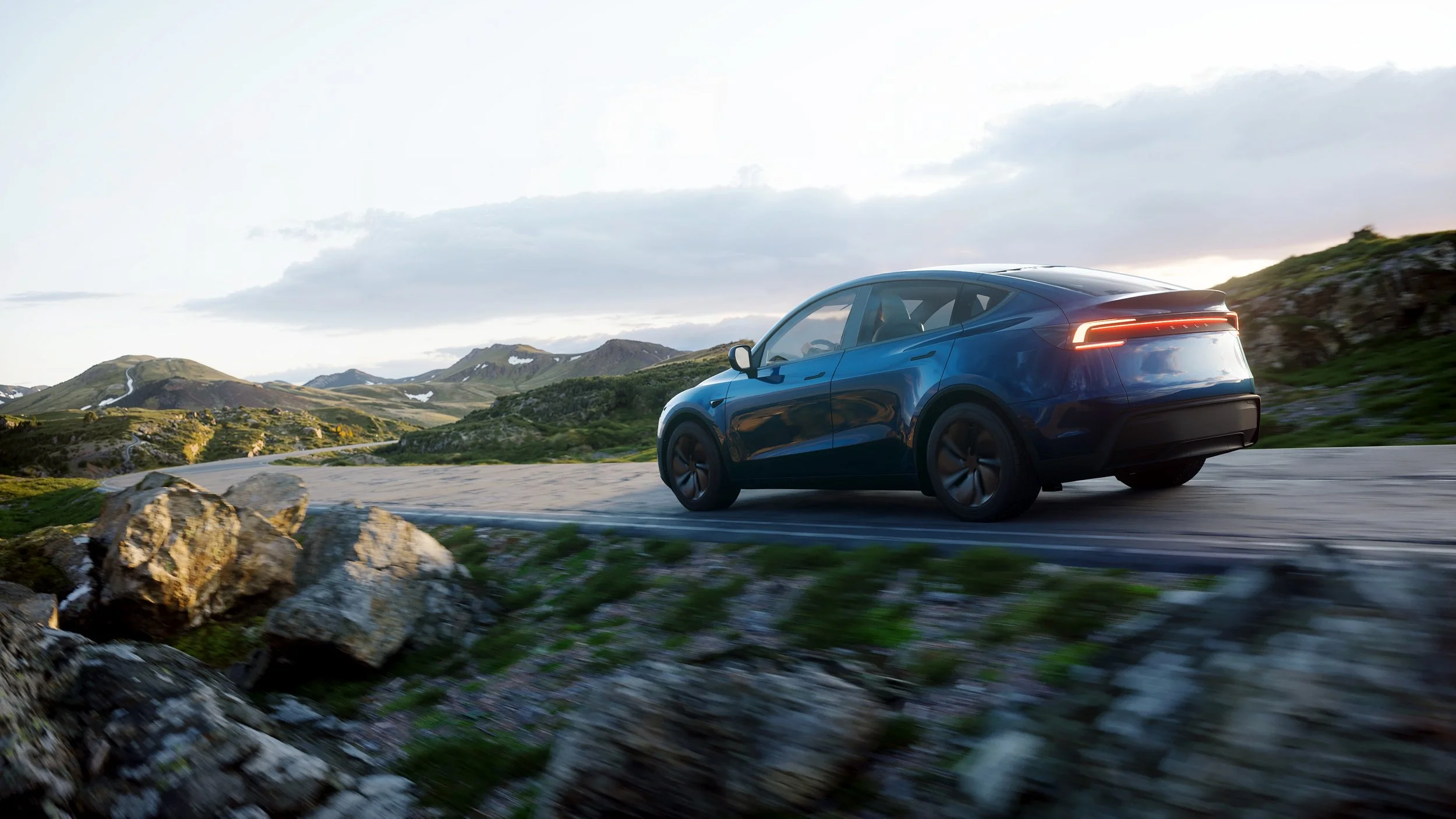 A blue electric vehicle driving on a winding mountain road surrounded by green hills and rocks, with a partly cloudy sky.