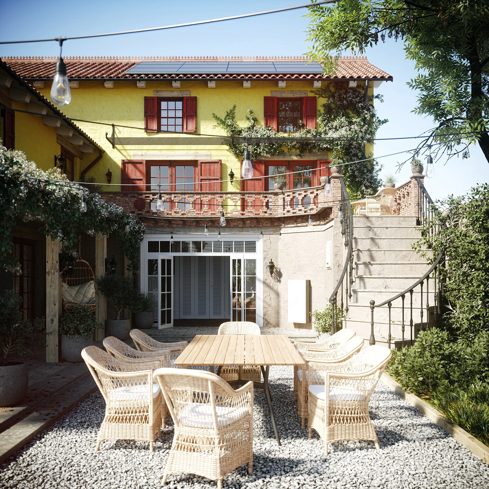 Outdoor patio area with a wooden table and wicker chairs on gravel, adjacent to a yellow house with red shutters and a balcony, surrounded by greenery and string lights.
