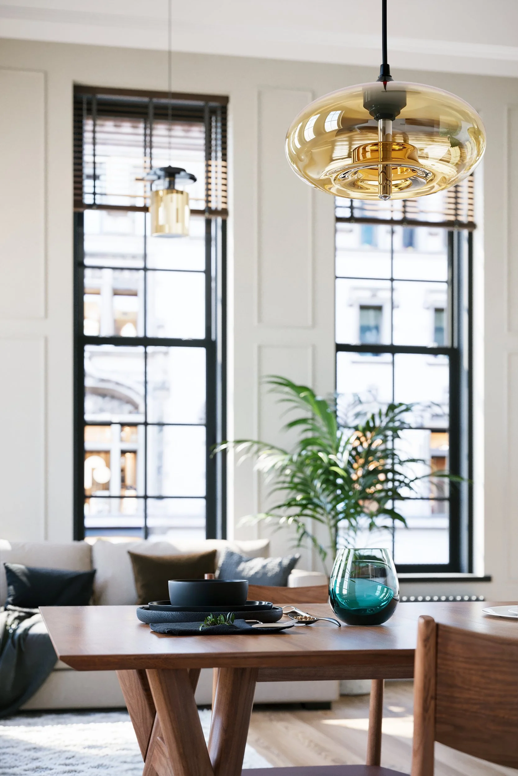 A modern living room with a wooden dining table set with black bowls, a teal glass vase, and spoons. In the background, two large windows with blinds, a light-colored sofa with pillows, and a potted plant near the window are visible. Hanging from the