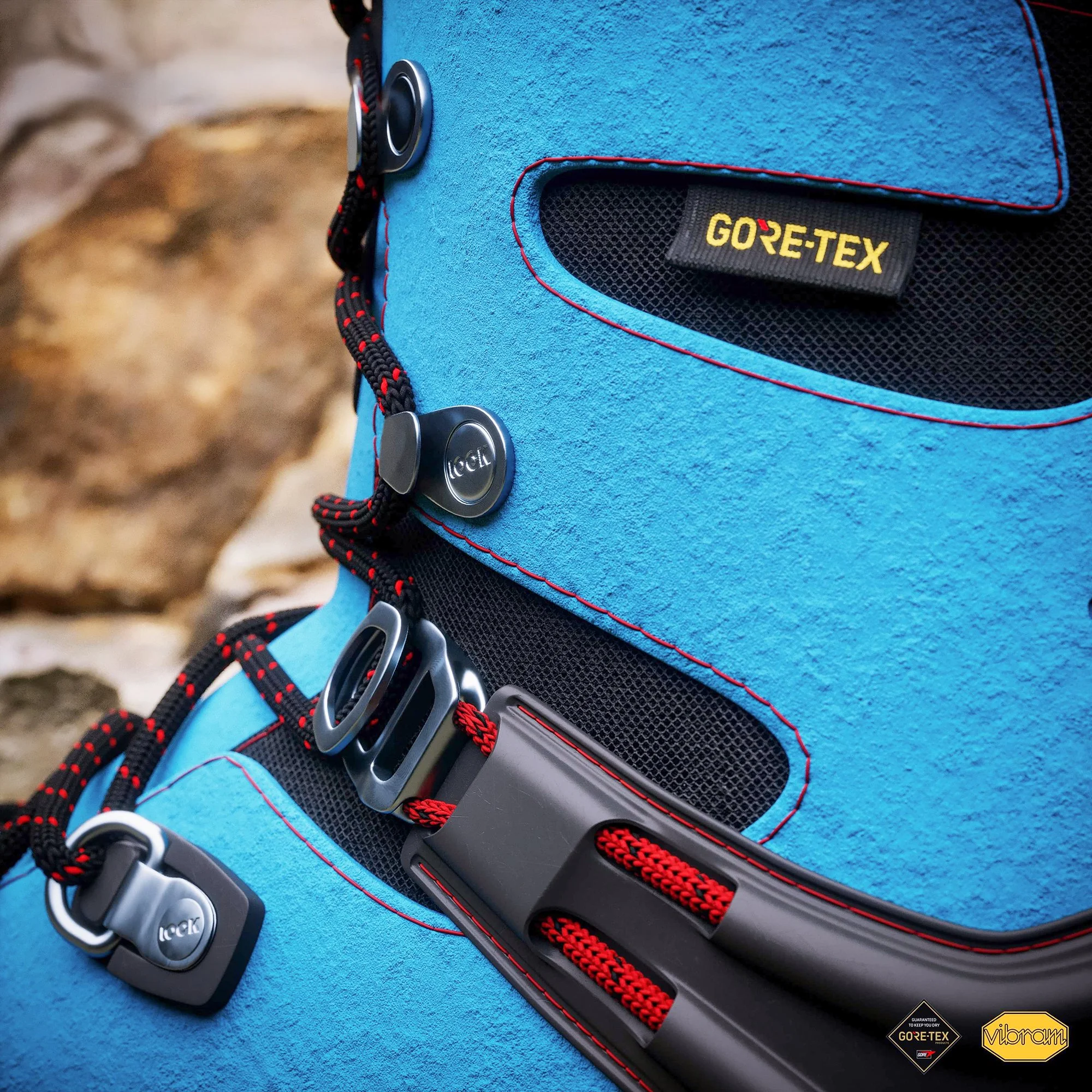 Close-up of a blue outdoor backpack with black mesh pockets, red stitching, and a black strap with red and black braided laces. The backpack features a GORE-TEX label and a Vibram logo at the bottom right corner.