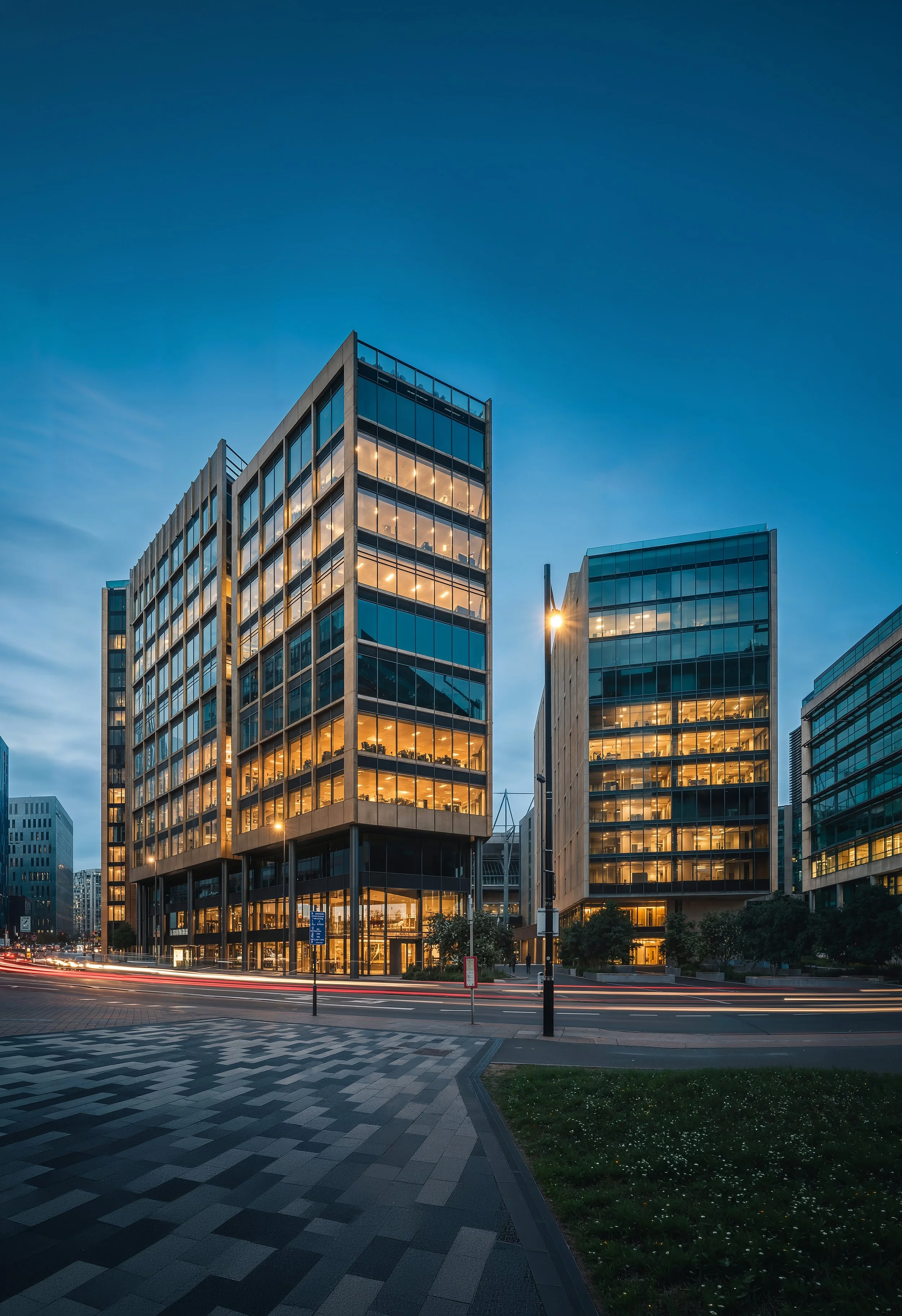 Modern office buildings with glass facades glowing with interior lights at dusk, on a city street with motion blur of traffic and a pedestrian crosswalk in foreground.