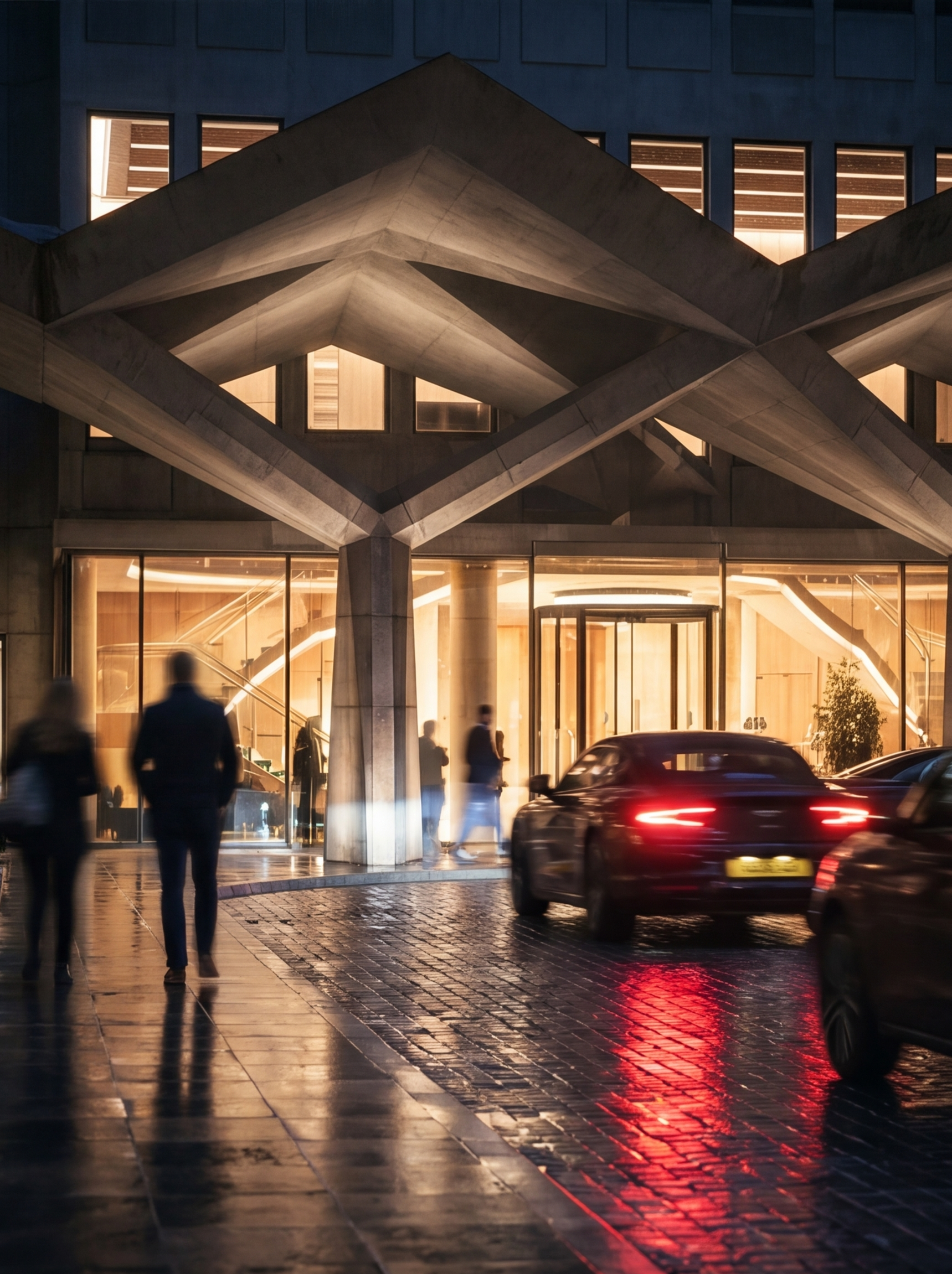 Night scene of a modern building with illuminated windows, people walking on wet pavement, and a car driving by, with reflections on the ground.
