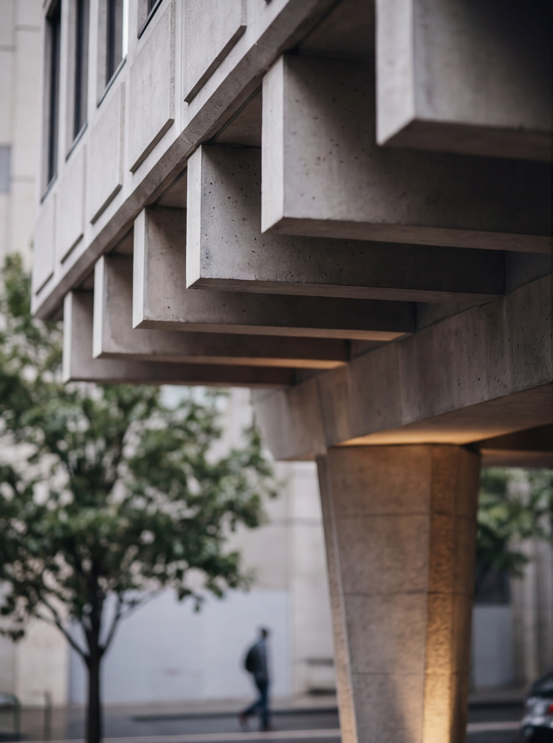 Close-up view of a modern concrete building with a concrete ledge and windows, blurred background showing a person walking and trees.