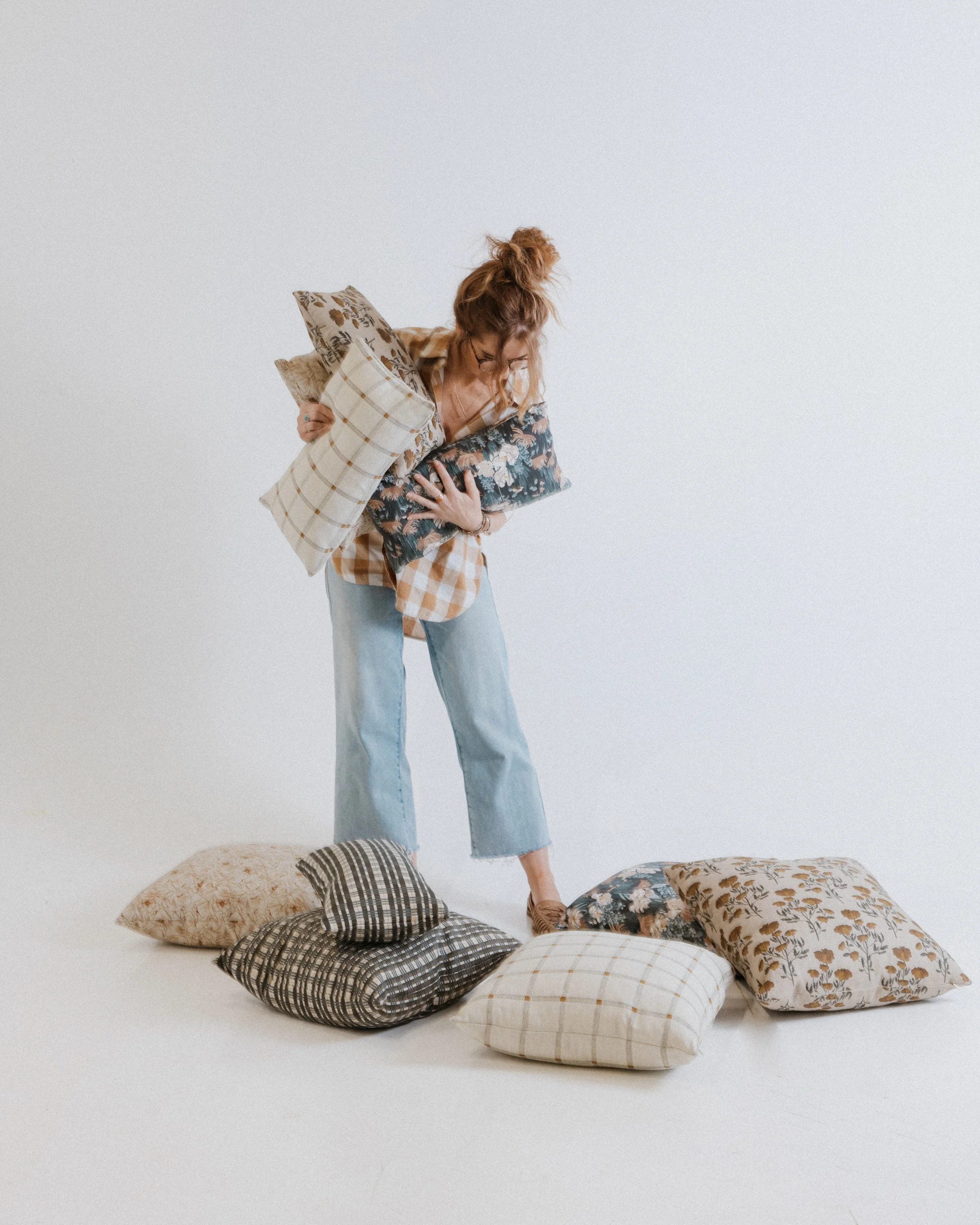 A woman with red hair, glasses, and casual clothing stands on pillows with floral and checkered patterns on the floor against a plain white background, holding multiple decorative pillows.