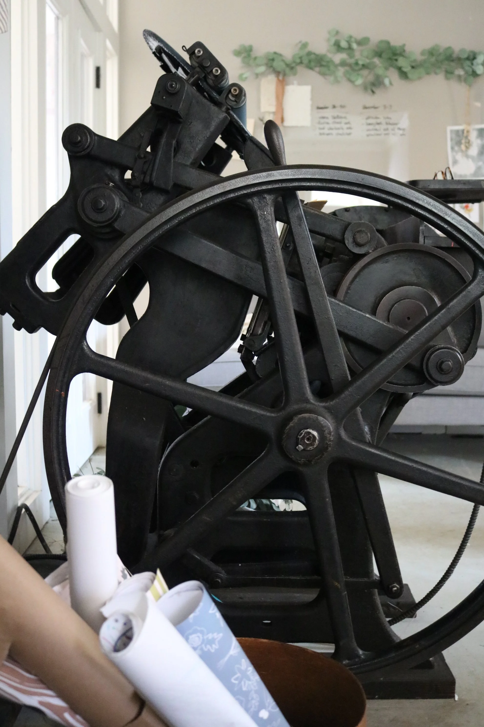 Close-up of a vintage black manual printing press or press machine with large wheels and gears in a room with light-colored walls and decorative greenery in the background.