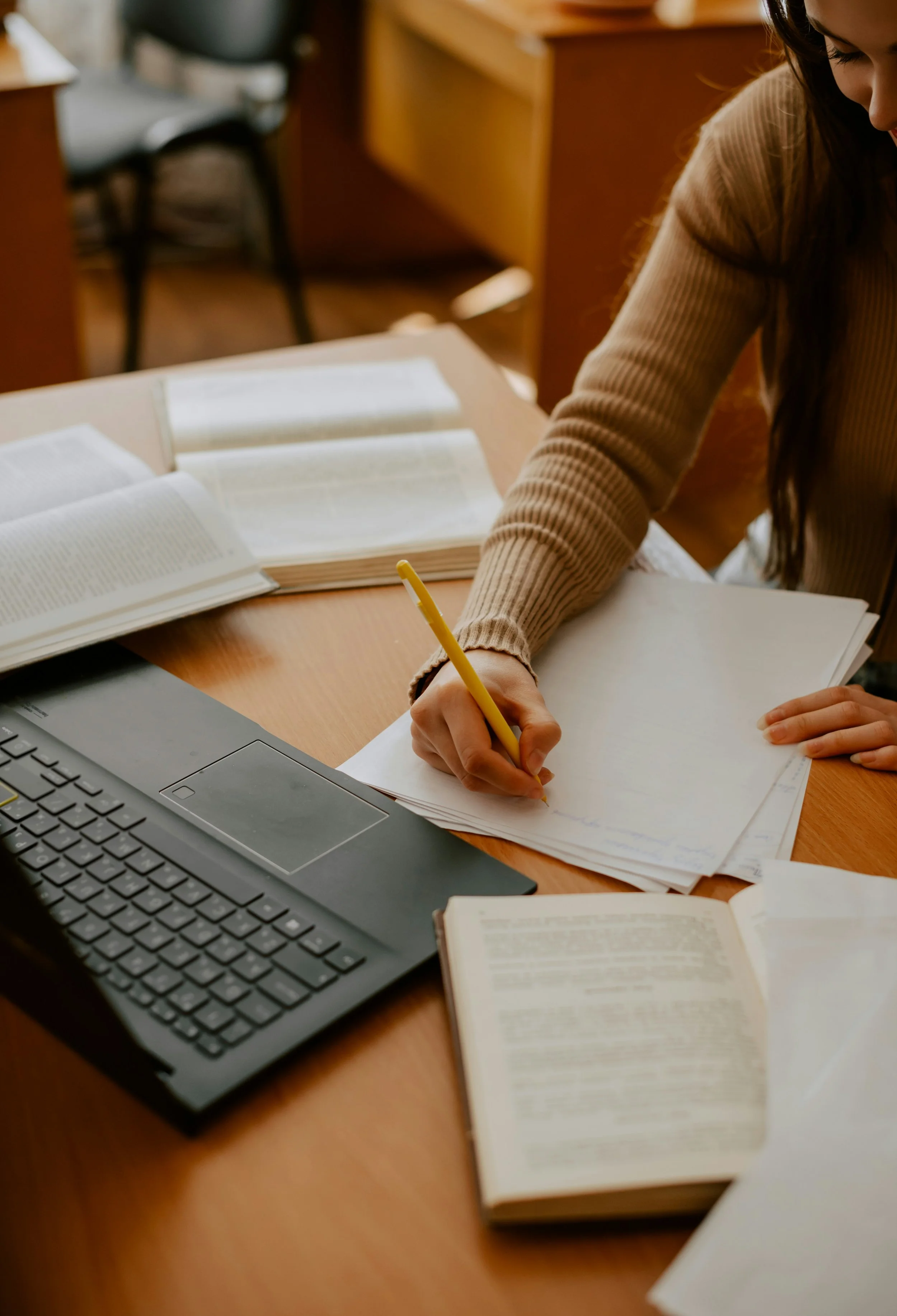 A person with long dark hair wearing a beige ribbed sweater is writing on a large sheet of paper with a yellow pencil at a wooden desk. There are open books, a laptop, and additional papers on the desk.