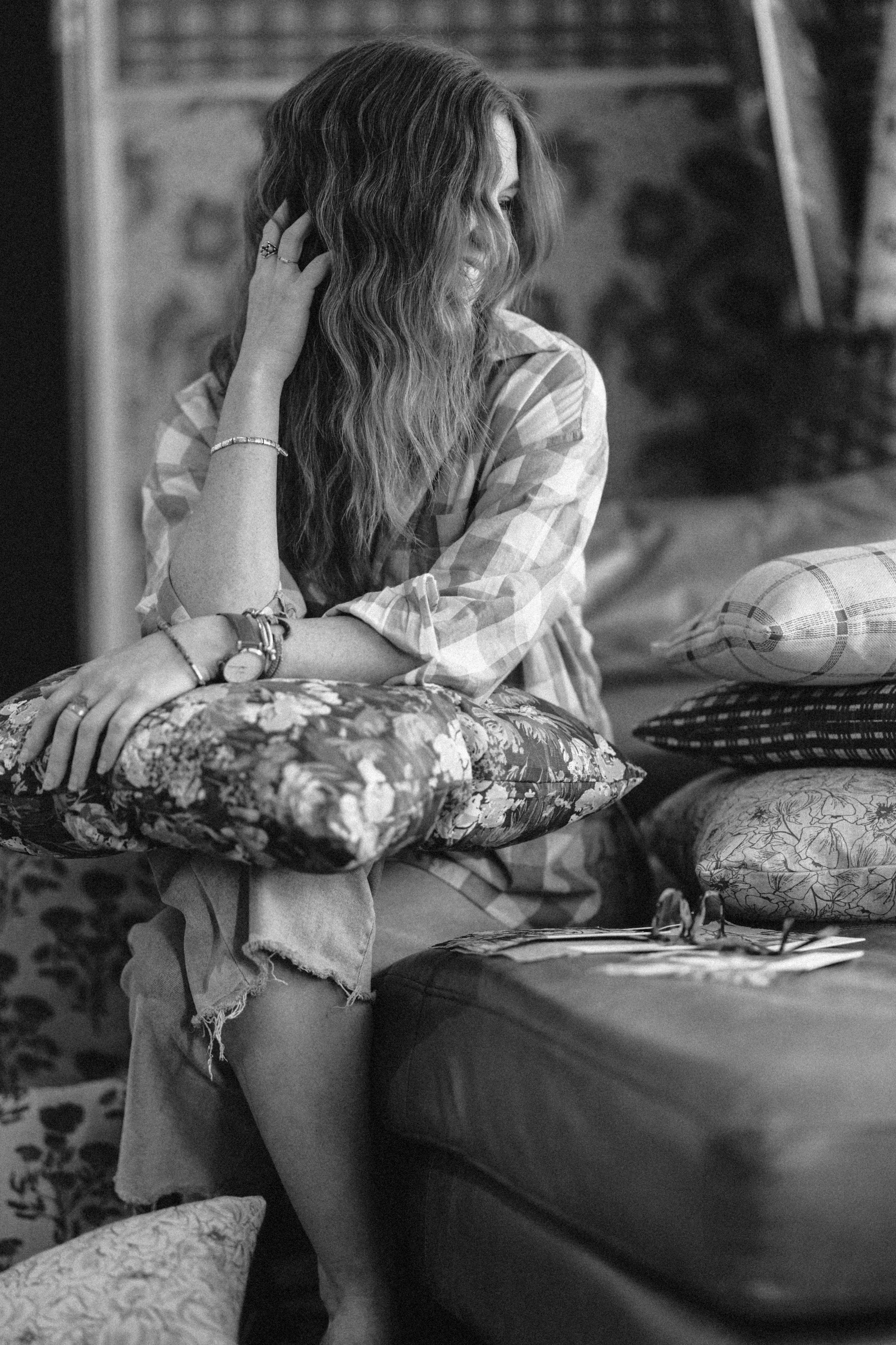 A woman with long wavy hair sitting on a cushioned surface, smiling and touching her hair, wearing a plaid shirt, necklace, and watch, with a background featuring plants and a rustic setting.