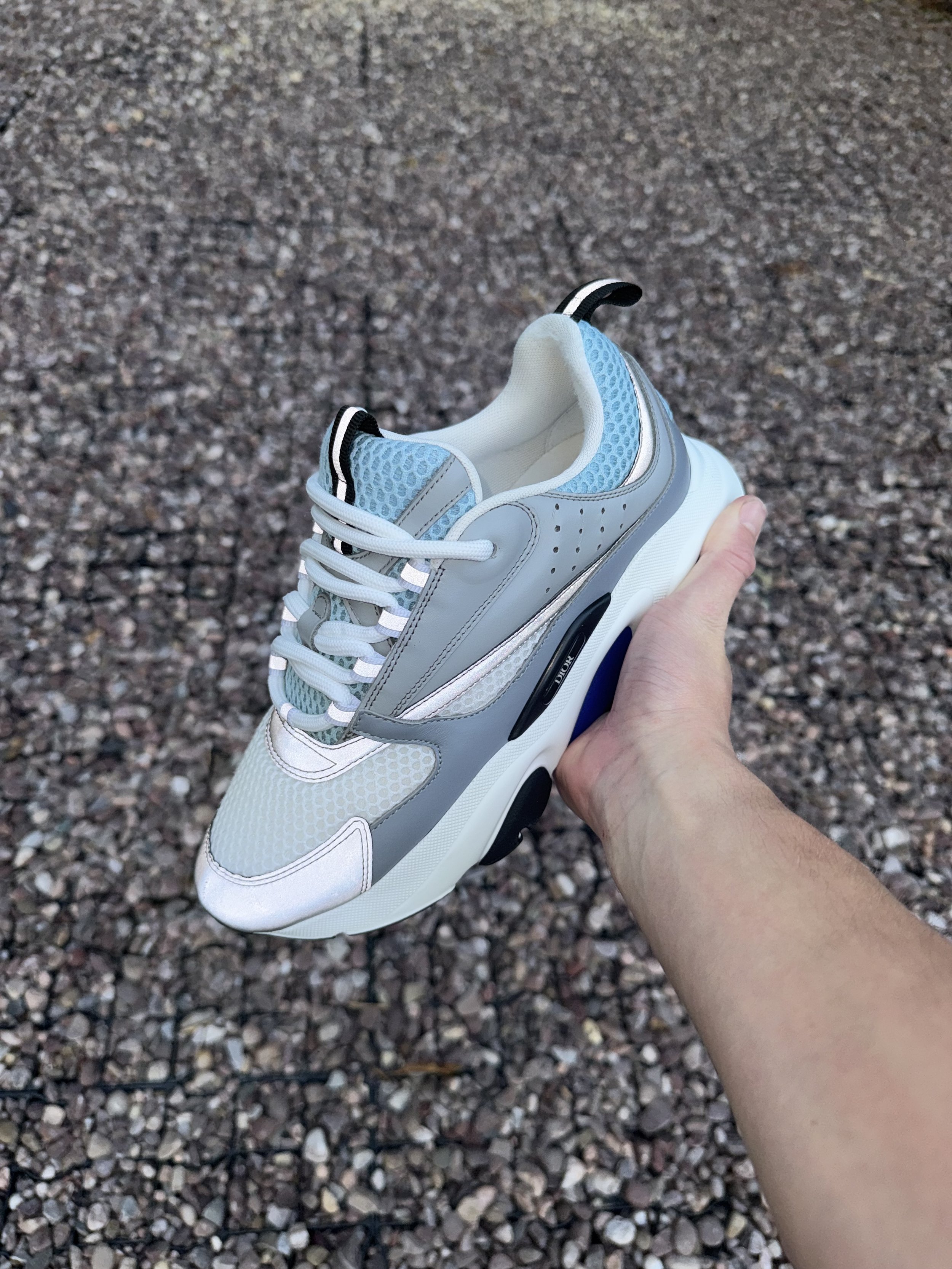 A person holding a stylish sneaker in their hand, with a gravel ground in the background.