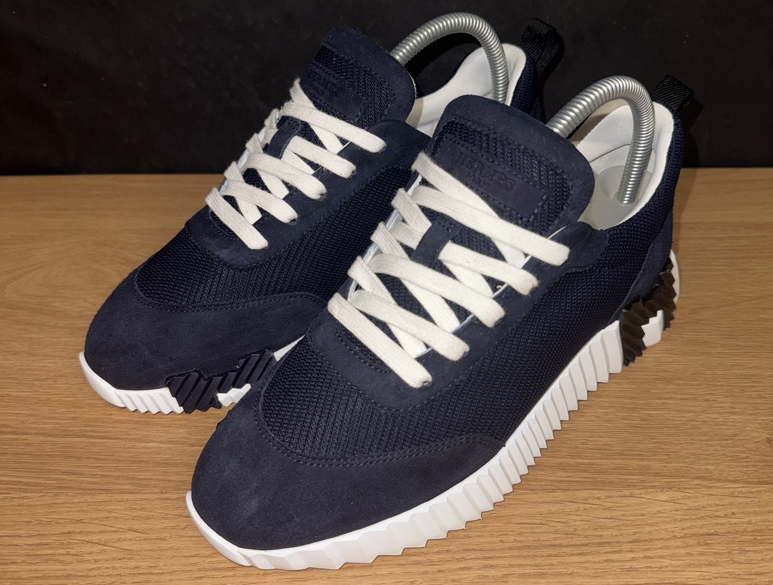 A pair of navy blue sneakers with white laces and a split sole, placed on a wooden surface with a black background. The sneakers have gray shoe horns inserted.