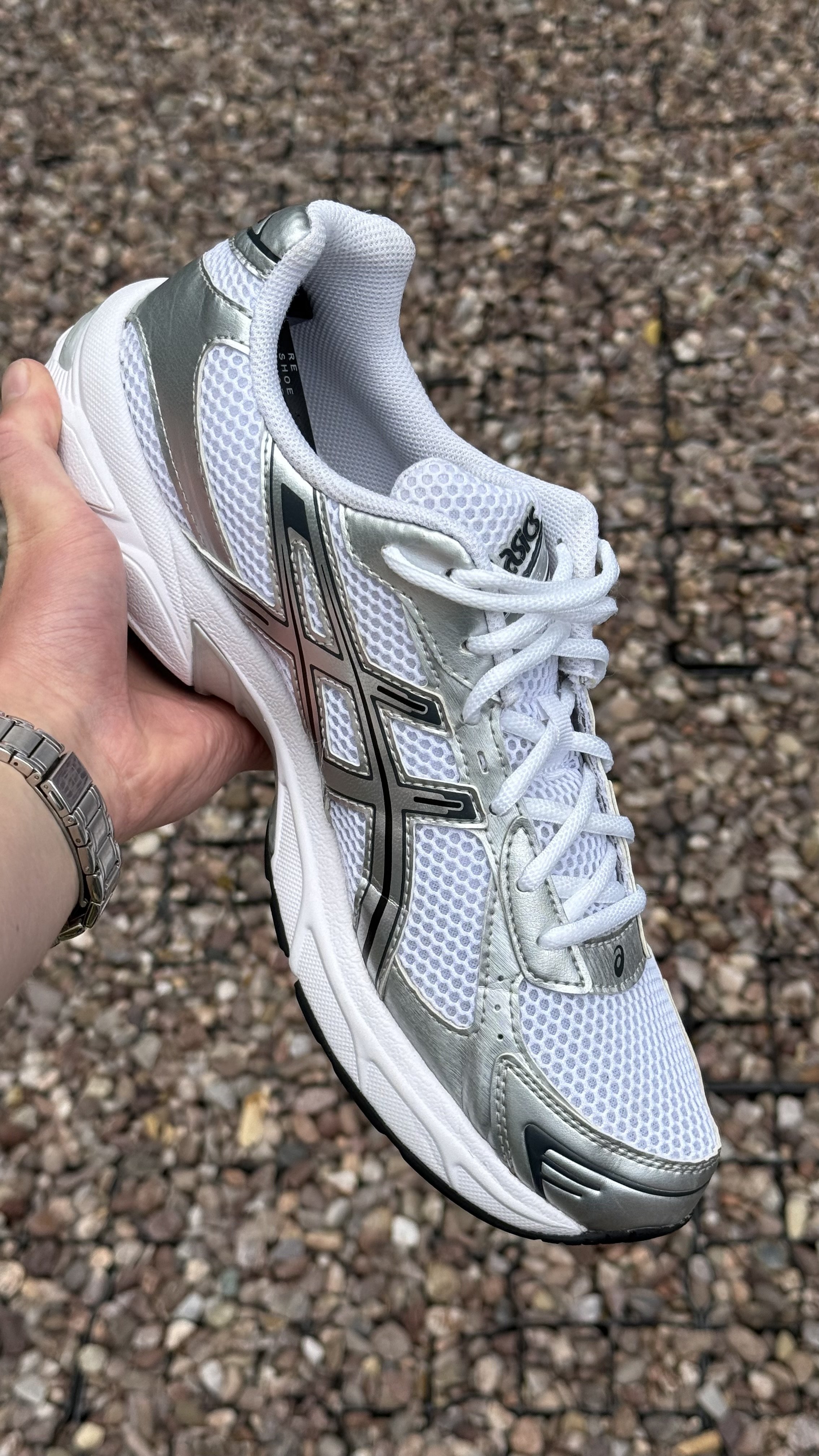 A white and silver athletic shoe with mesh and synthetic overlays, held by a person's hand, with a background of small rocks or gravel.