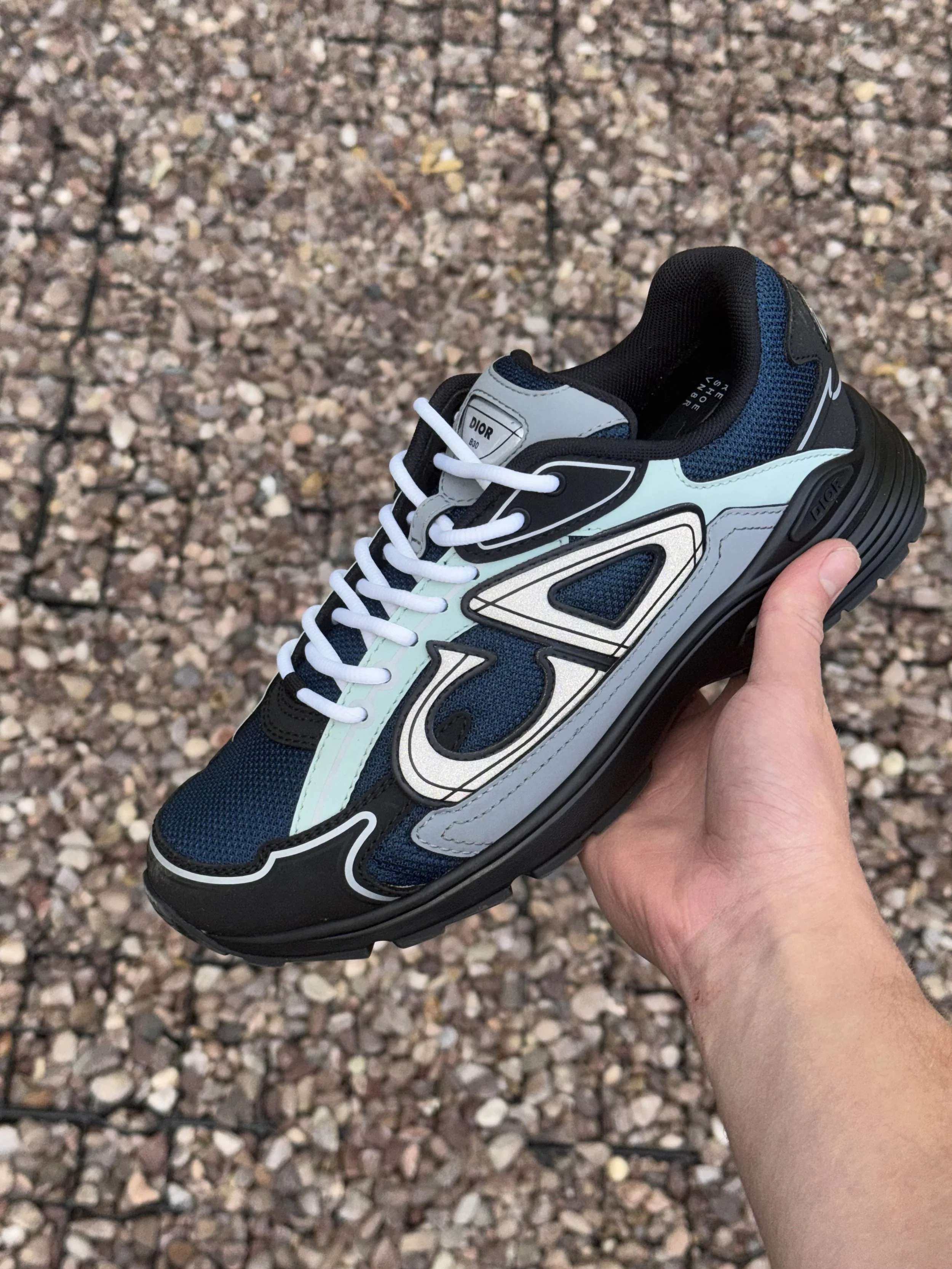 A close-up of a sneaker with blue and white details, held in a person's hand against a rocky ground background.