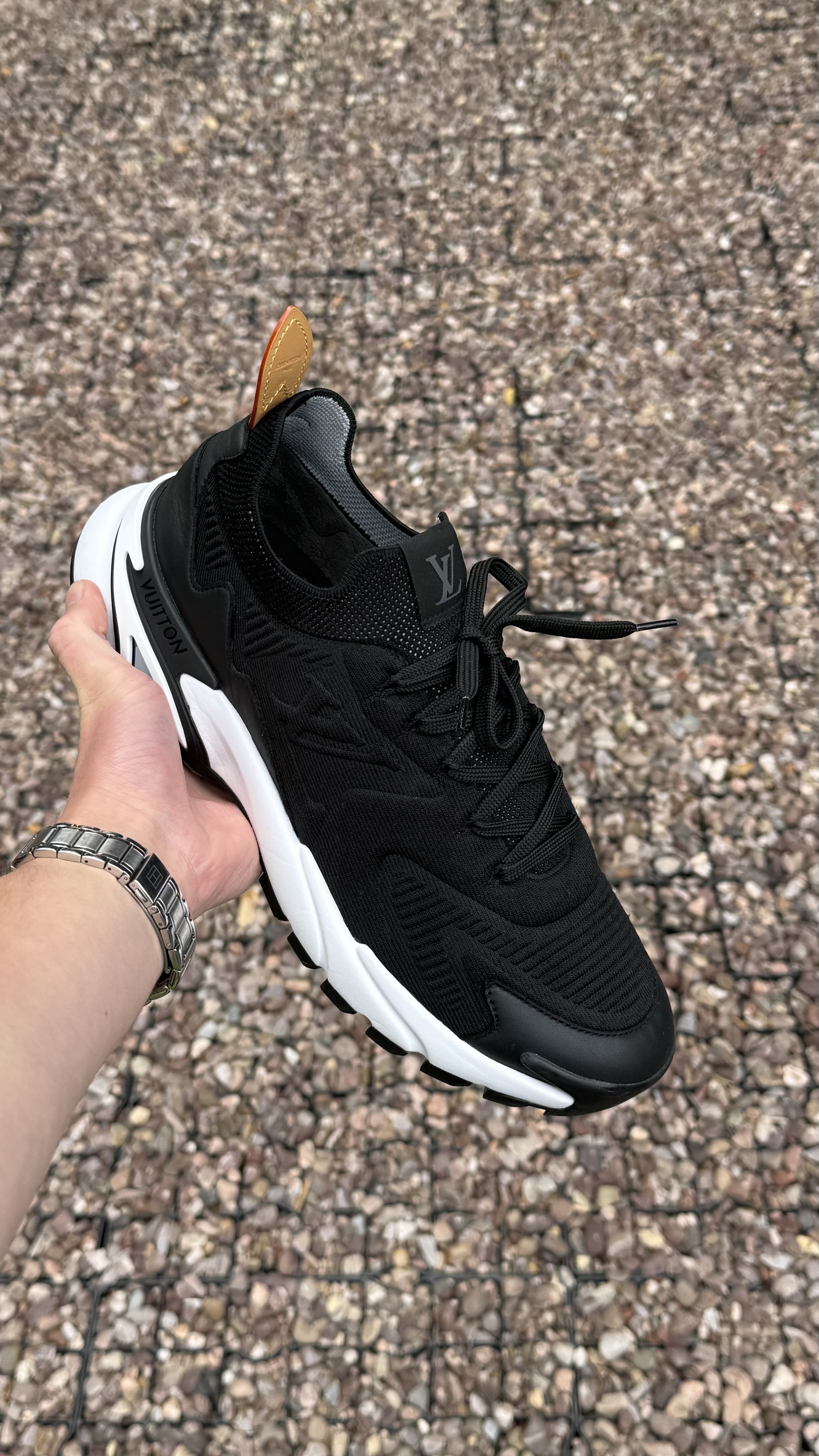 Hand holding a black Adidas Yeezy sneaker with a white sole, on a gravel ground.