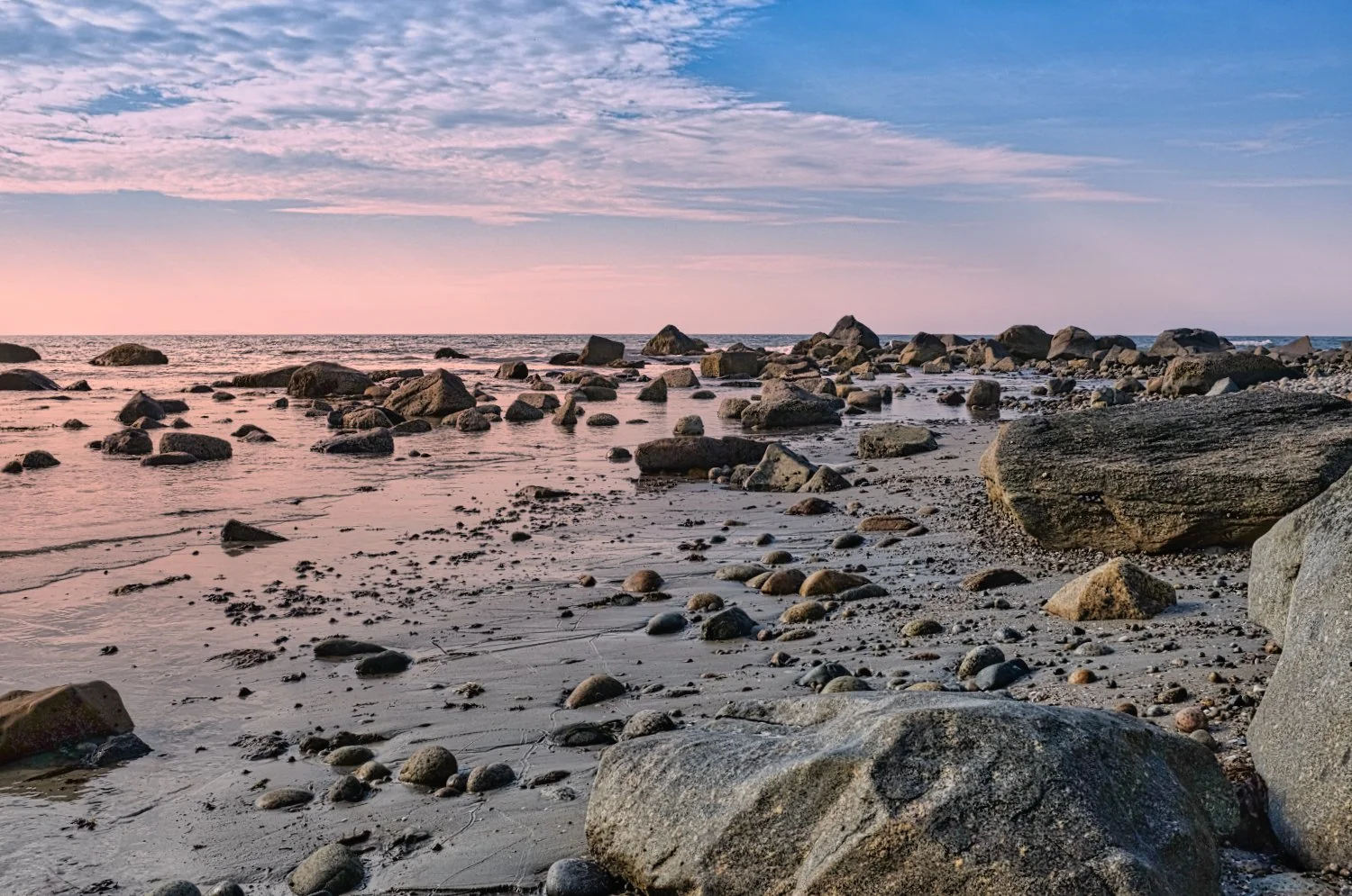 Sunset over a rocky beach with scattered stones and calm waves, pink and blue sky with some clouds.
