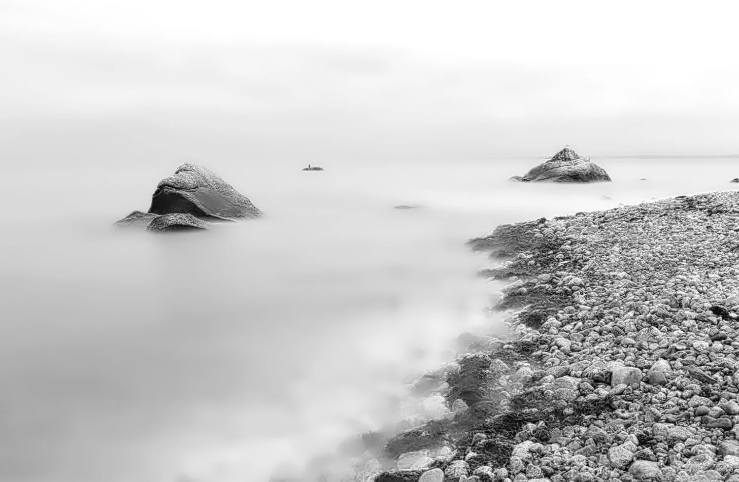 Black and white photo of a rocky shoreline with large rocks extending into calm water, smooth due to long exposure, with a distant small rock formation with a lighthouse on top, and a cloudy sky.