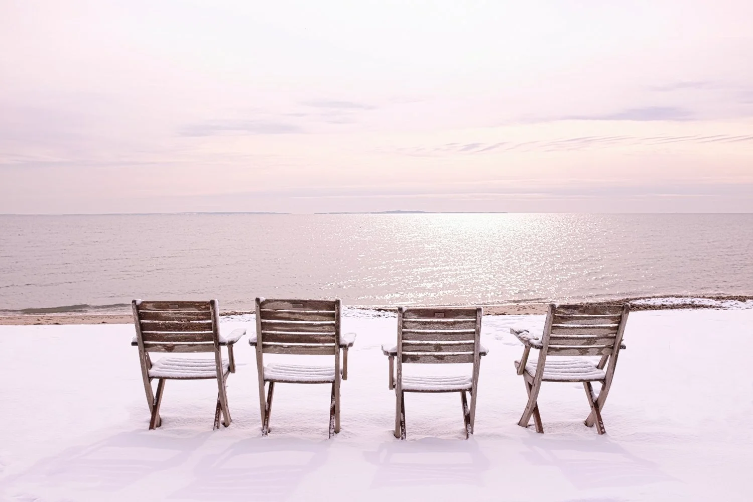 Four wooden chairs covered in snow facing a calm, icy ocean under a gray sky.
