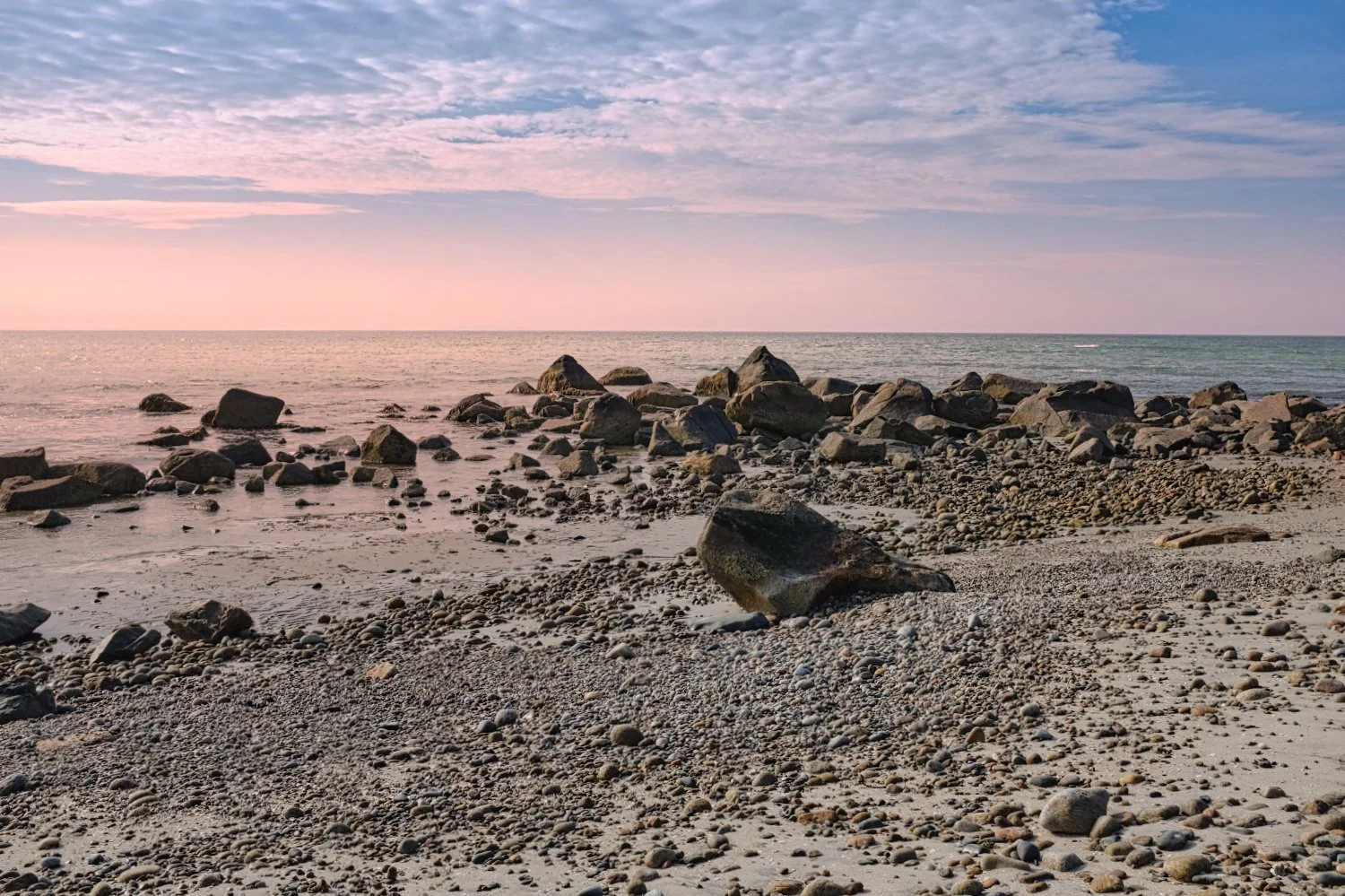 A rocky beach scene at sunset with a pastel pink and blue sky, scattered rocks and pebbles on the shore, and gentle ocean waves in the background.