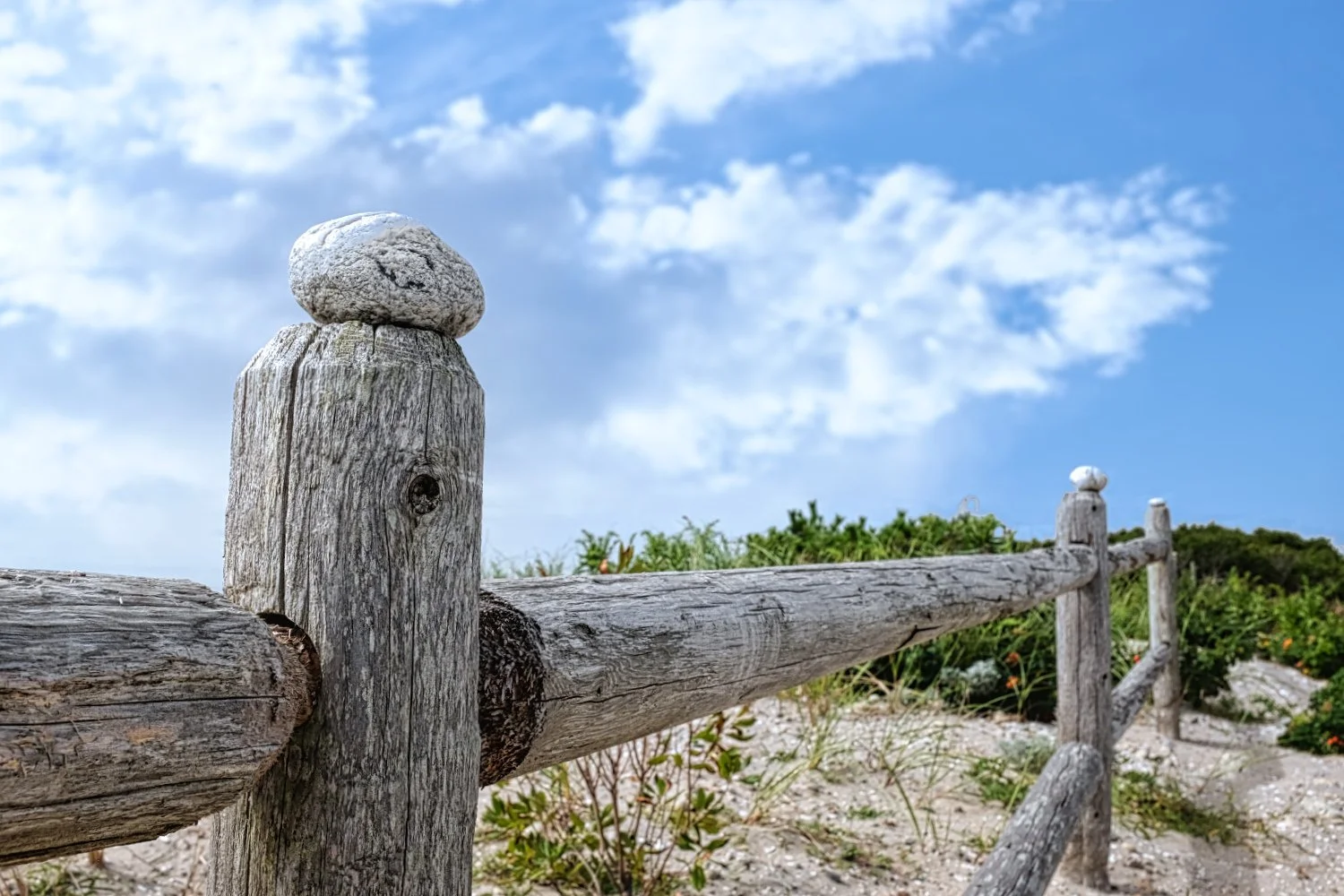 A weathered wooden fence with a round stone on top of a post, set against a backdrop of green shrubbery, sandy ground, and a partly cloudy blue sky.