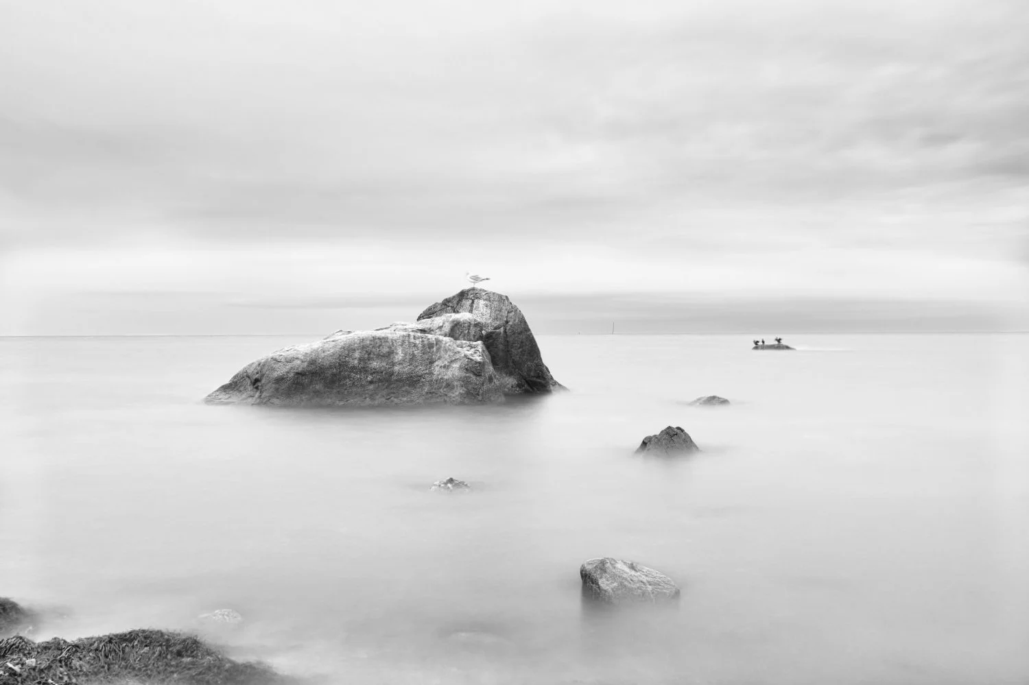 Black and white photo of large rocks in calm water with a seagull perched on one rock and a boat with two people in the distance under a cloudy sky.