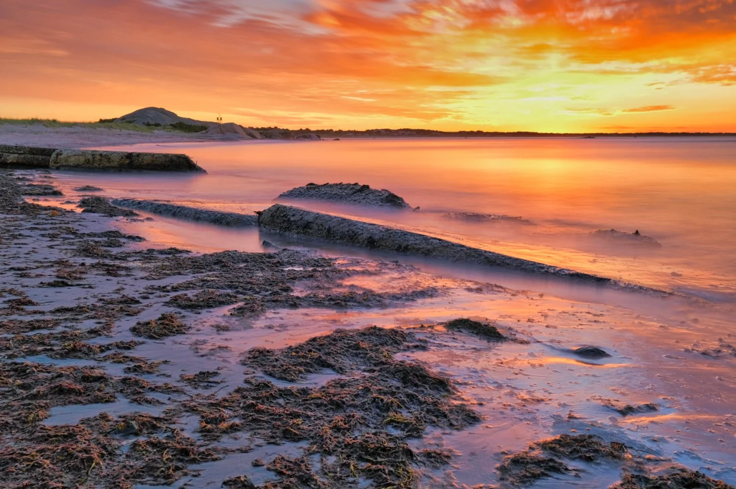 Sunset over a beach with rocks, sand, and calm water, with orange and yellow sky reflected on the water
