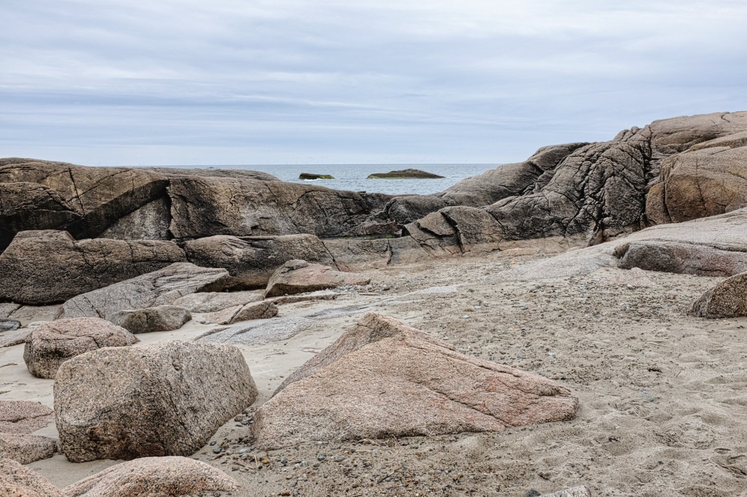 A rocky beach with large granite stones and sandy patches, overlooking the ocean with a cloudy sky.