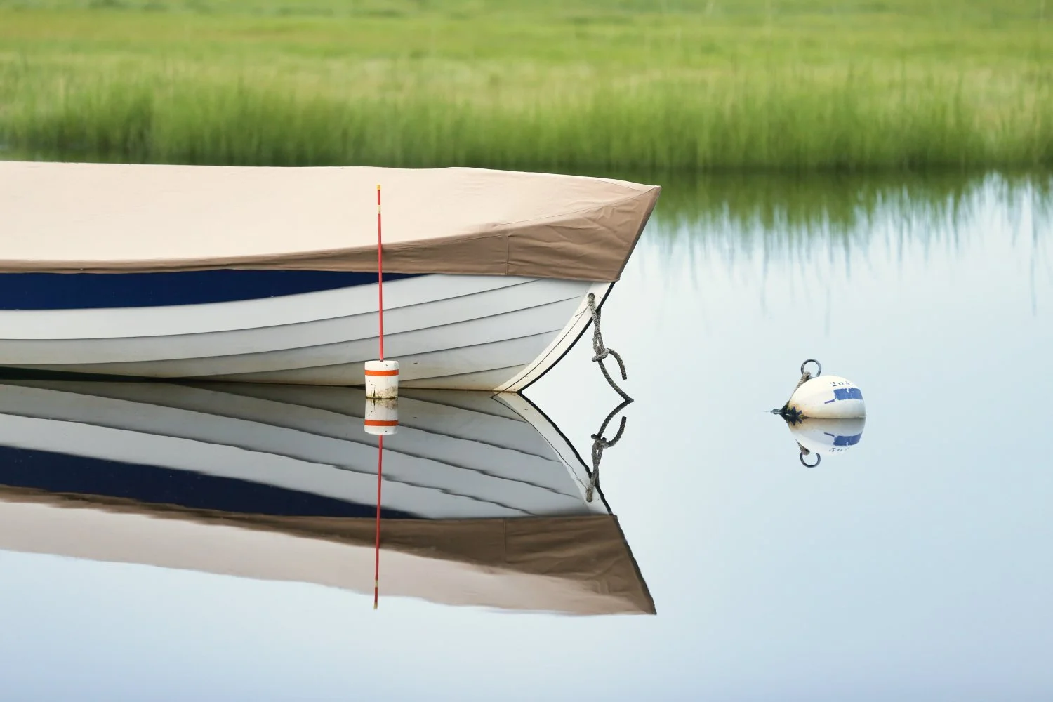 A boat moored in calm water with a reflection of the boat and a buoy attached to it; the background shows green grasses and trees.