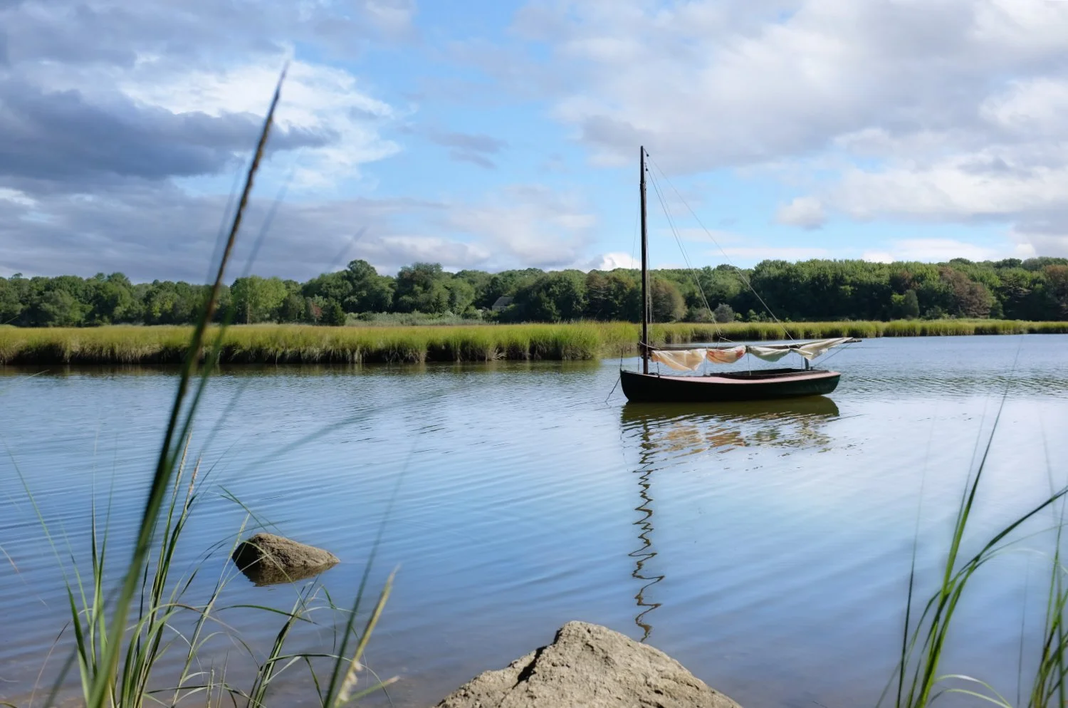 A sailboat floating on a calm river with a grassy shoreline and lush trees in the background under a partly cloudy sky.