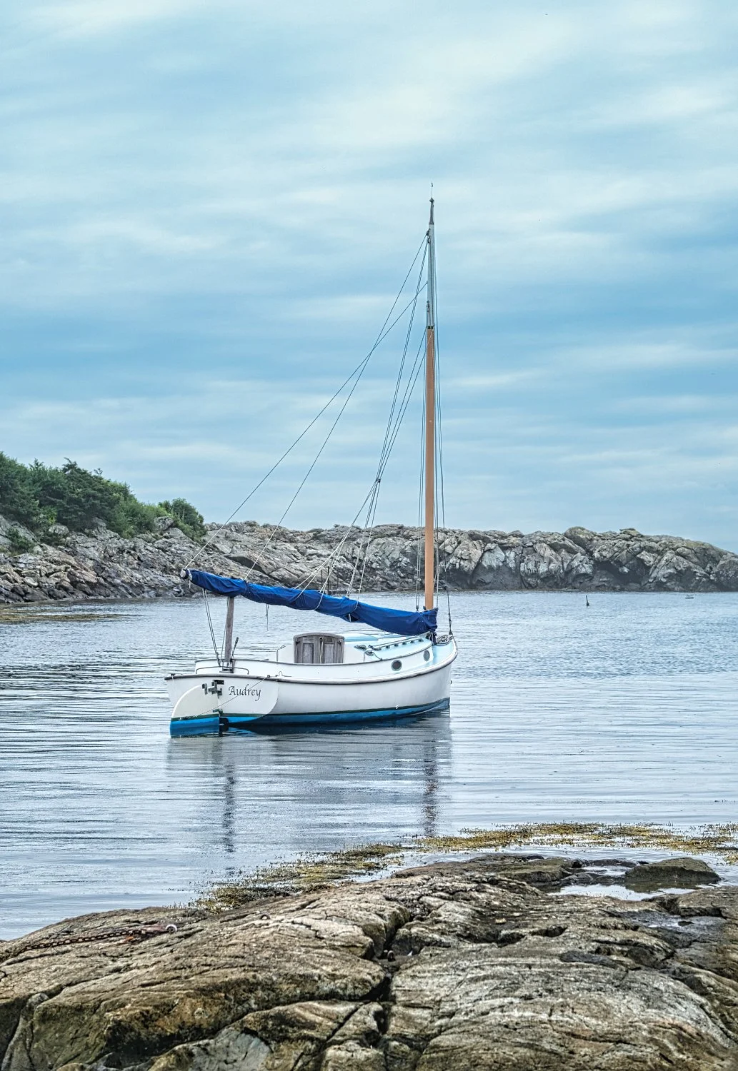 A sailboat named Audrey floating on calm water near rocky shoreline under cloudy sky.